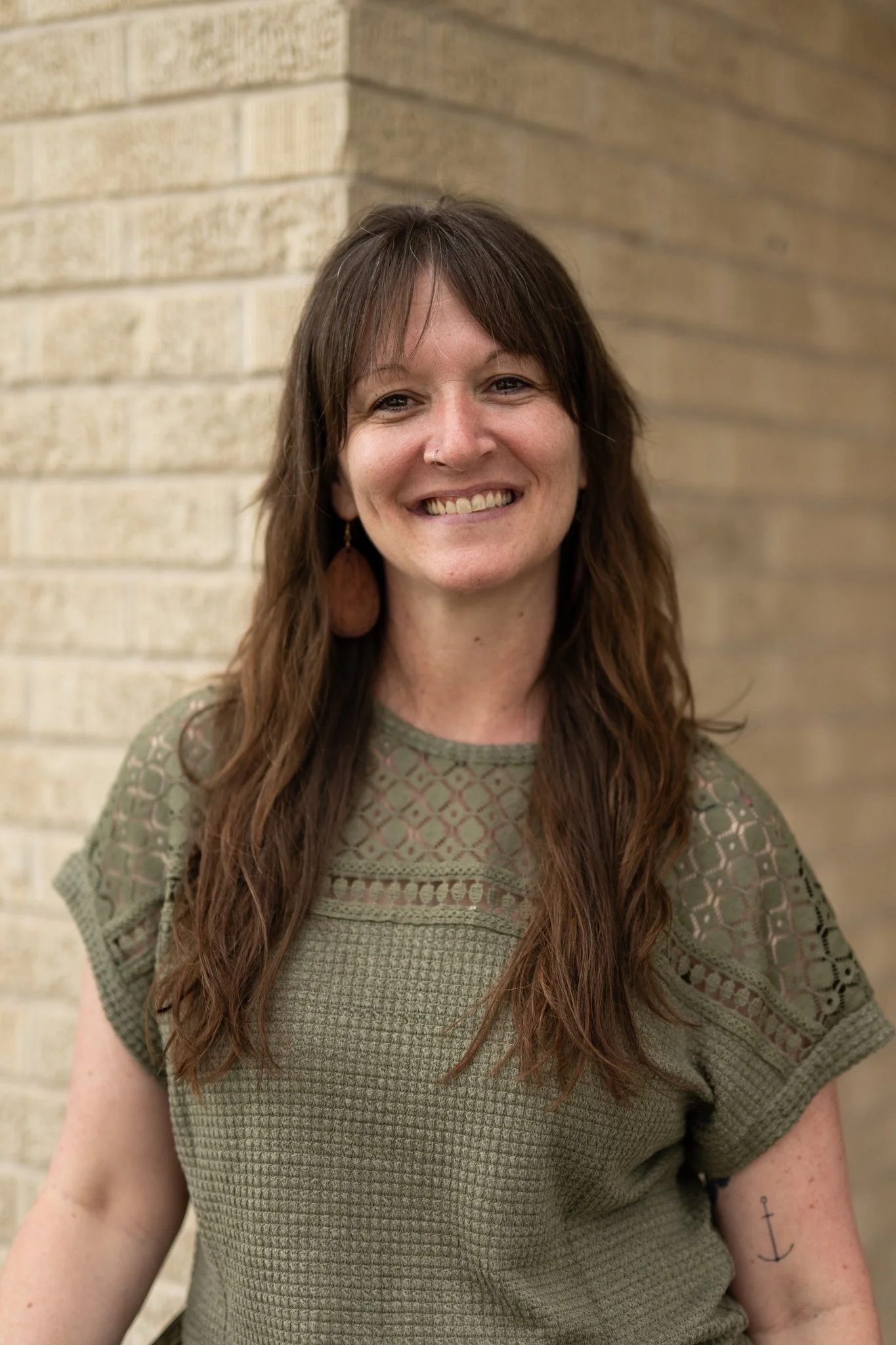 A woman with long brown hair wearing a green patterned top, smiling, standing in front of a beige brick wall.