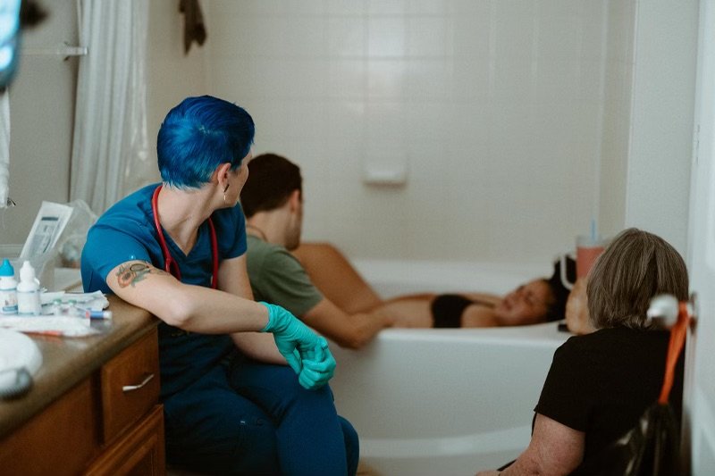 A nurse with blue hair and gloves sitting next to a man in a recovery bed, with a woman sitting nearby, in a hospital or clinic room.