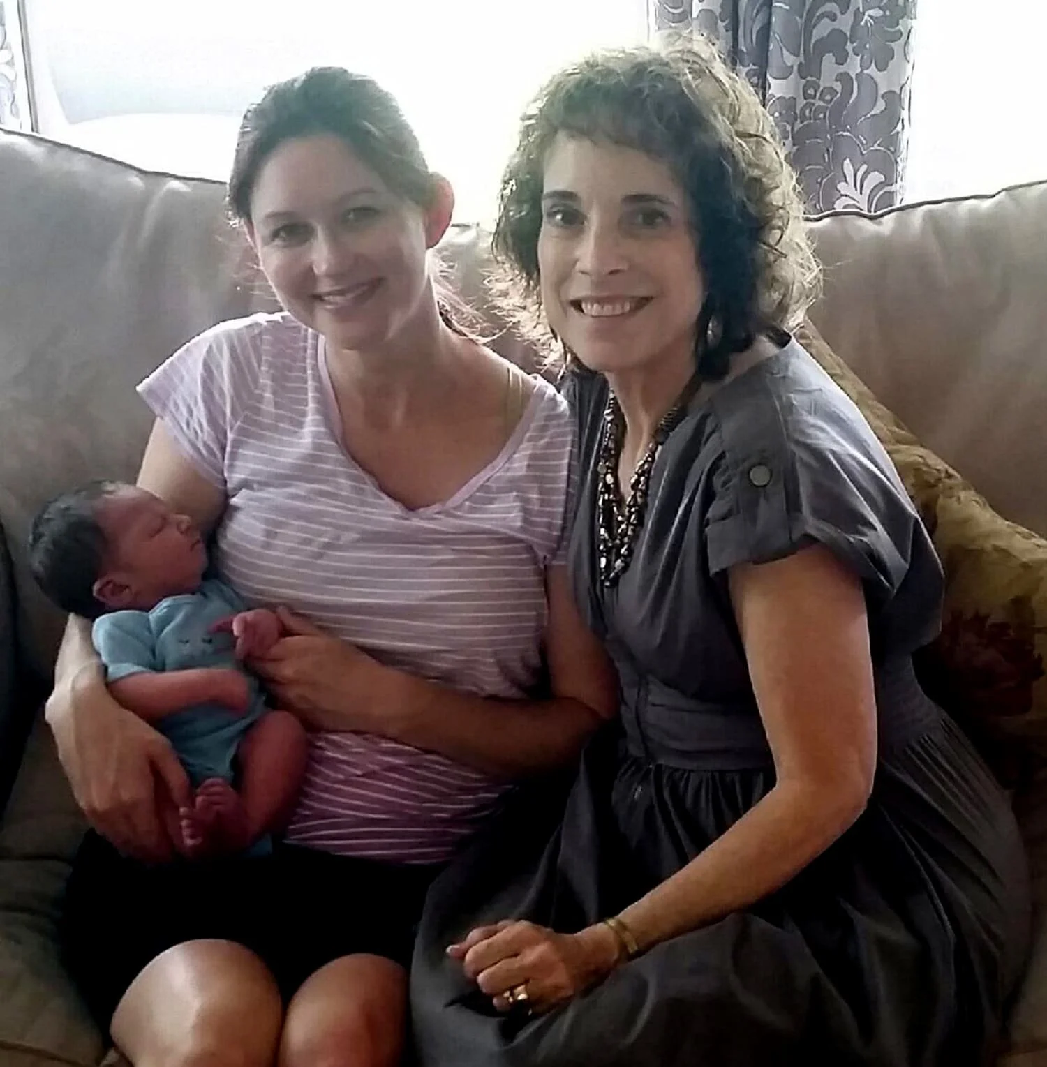Two women sitting on a couch, holding a newborn baby. The woman on the left has dark hair and is wearing a striped pink and white shirt. The woman on the right has curly dark hair and is wearing a dark gray dress with a necklace. The baby is dressed in a light blue outfit and is being held gently by the woman on the left.