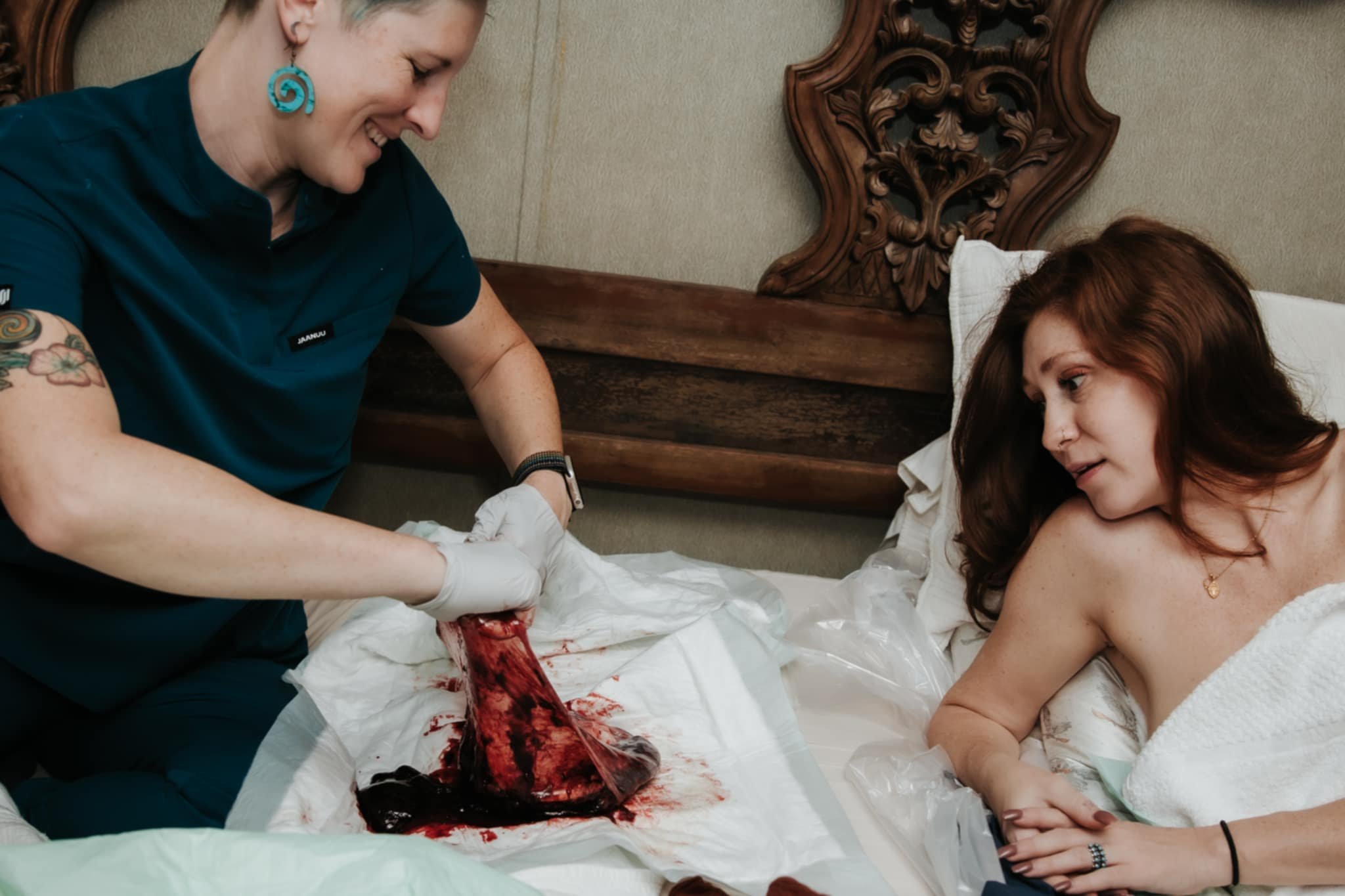 A woman with short gray hair and tattoos on her arm, wearing blue scrubs and gloves, is attending to a woman with auburn hair, lying in a hospital bed. The woman in bed appears to be giving birth, with blood and tissue visible, and smiling at the med