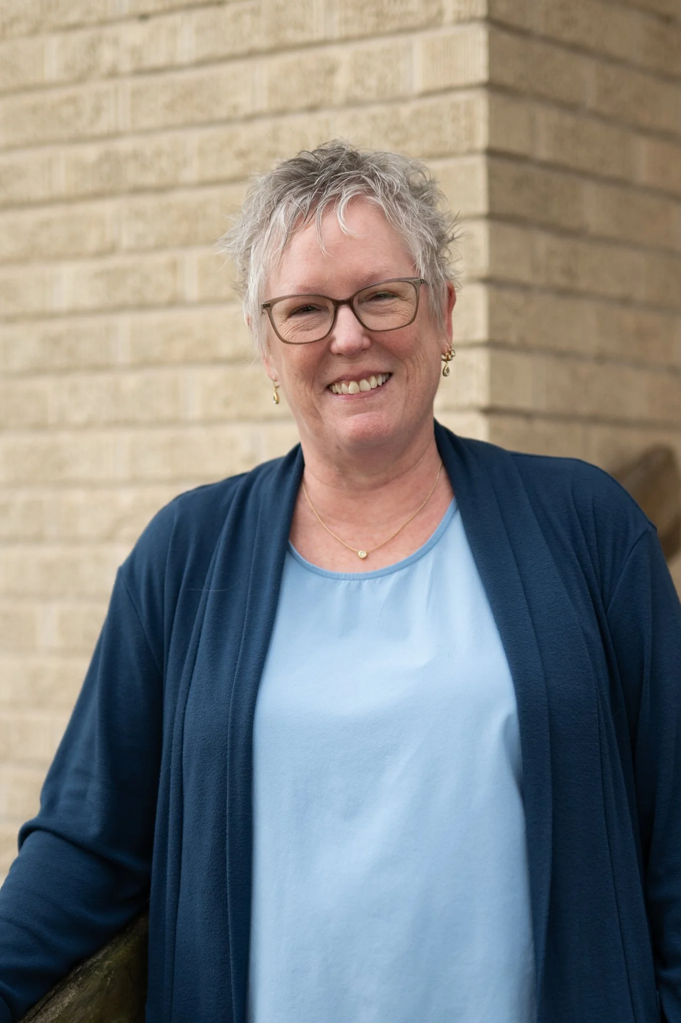 A smiling woman with short gray hair, wearing glasses, a gold necklace, gold earrings, a light blue shirt, and a navy blue cardigan standing in front of a beige brick wall.