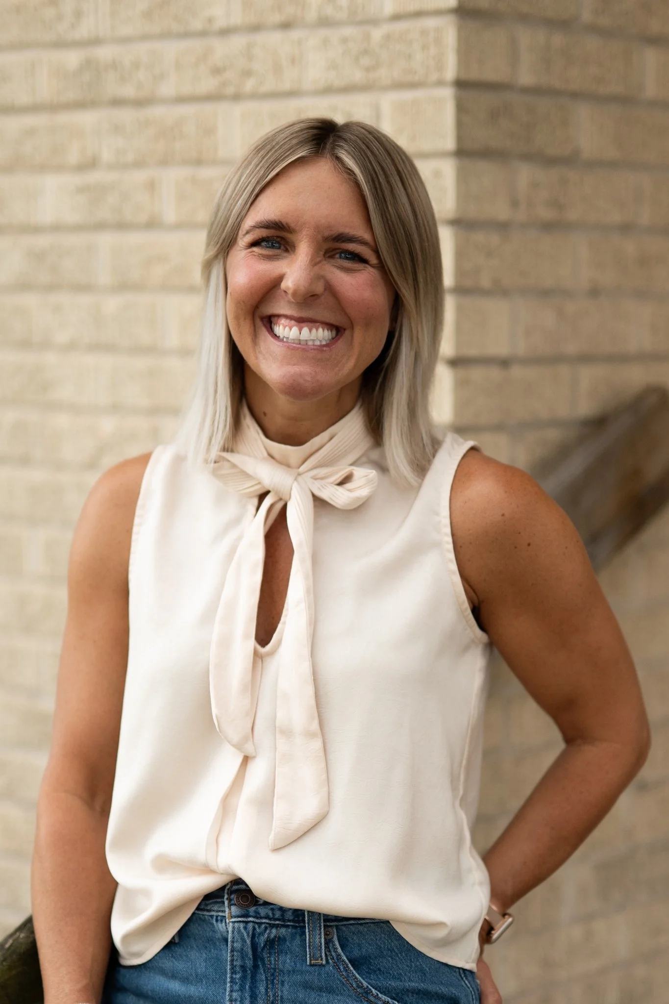A woman with blonde hair and blue eyes smiling, wearing a white sleeveless top with a bow tie at the neck and blue jeans, standing in front of a beige brick wall.