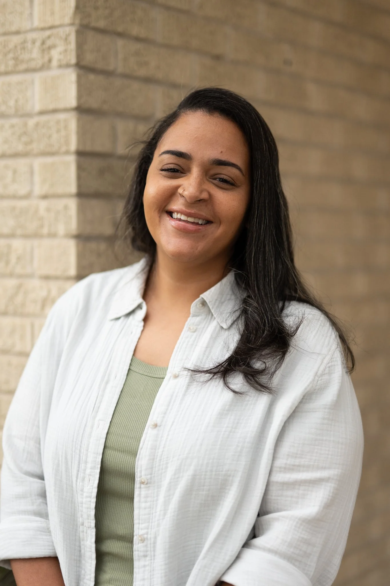 A woman with long dark hair smiling, wearing a light green top and a white striped shirt, standing against a brick wall.