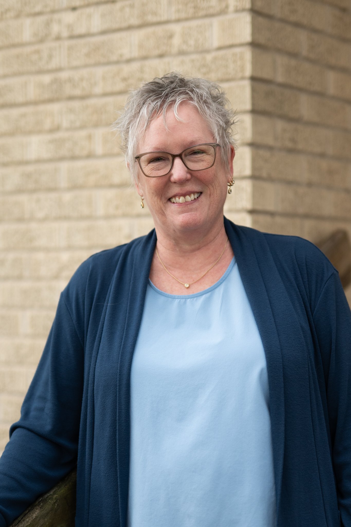 A smiling middle-aged woman with short gray hair, glasses, and dangle earrings, wearing a light blue top and a navy blue cardigan, standing outdoors against a beige brick wall.