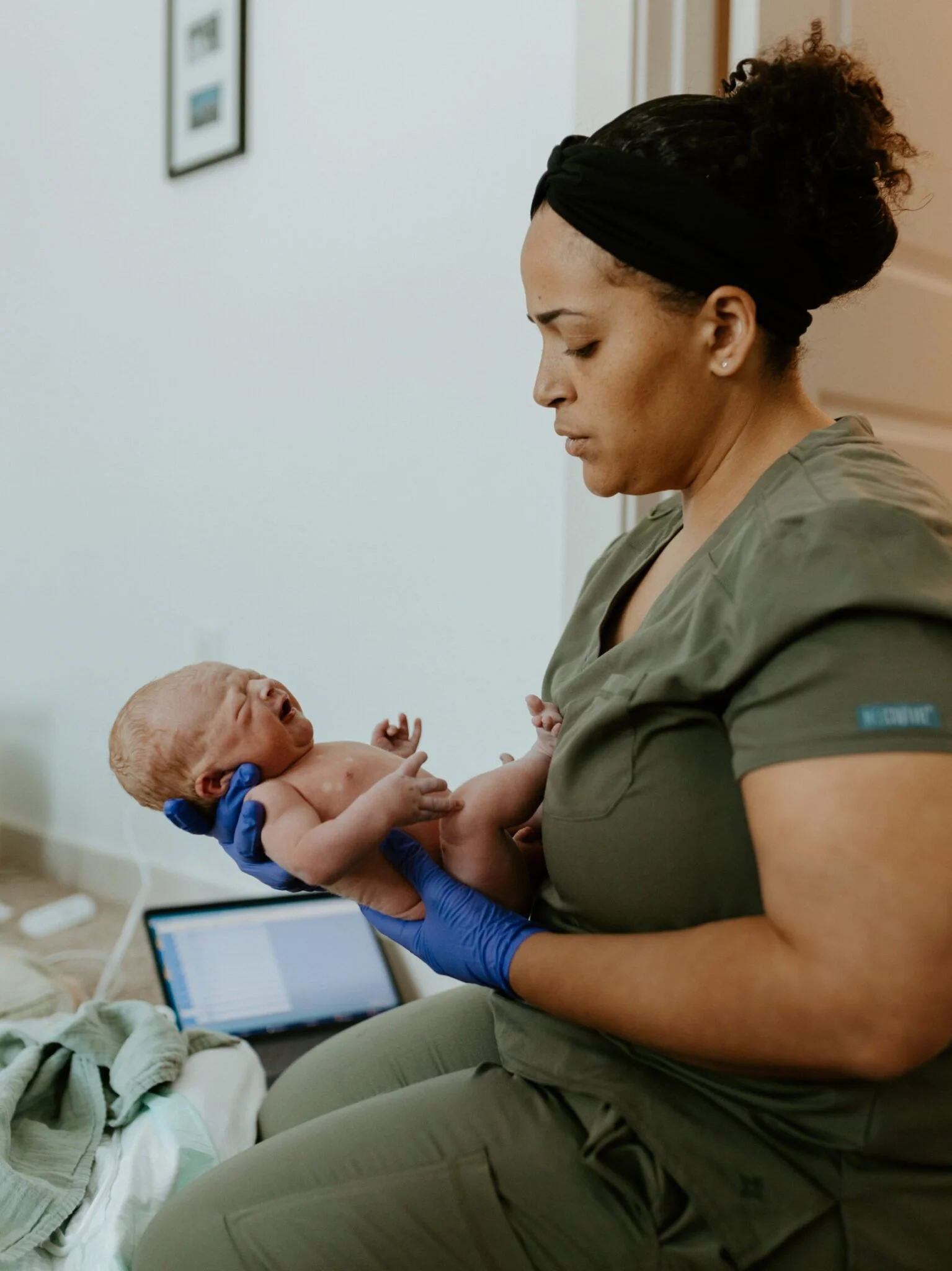 A woman, wearing green scrubs and a black headband, holding a fragile newborn baby with curly hair, inside Katy Birth Center.