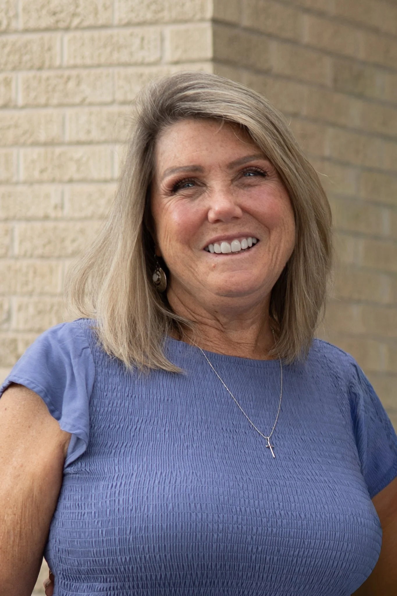 A woman with blonde hair smiling, wearing a blue top, earrings, and a necklace with a cross pendant, standing in front of a brick wall.