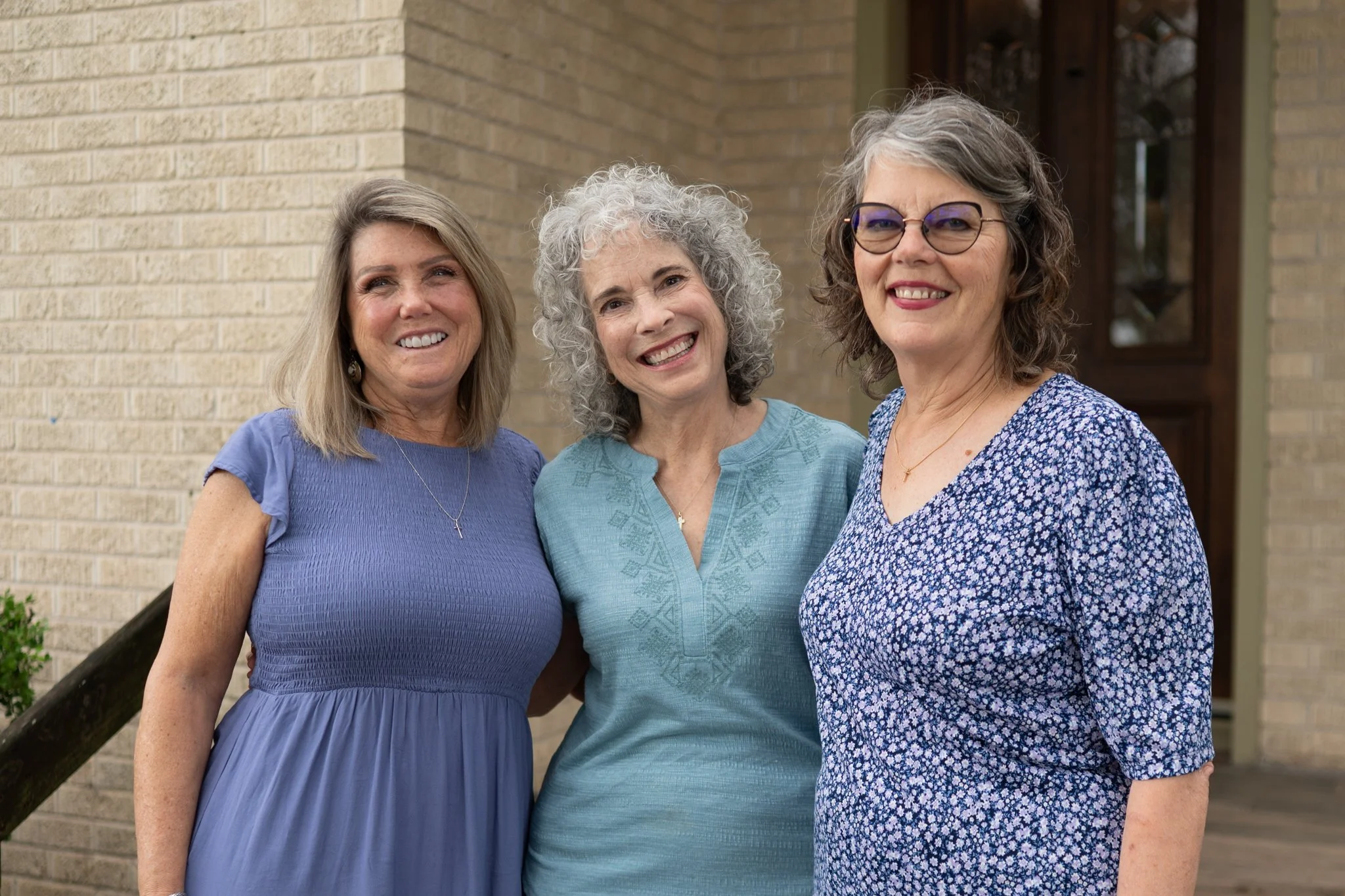 Three middle-aged women standing close to each other outdoors, smiling. They are dressed in blue tops and are standing in front of a brick wall with a window.