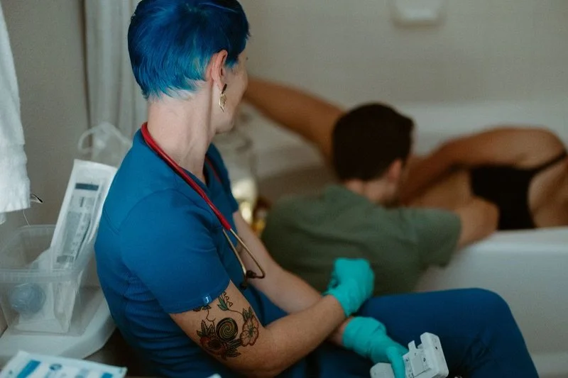 A nurse with short blue hair, tattoos on her arm, and a stethoscope around her neck, is sitting beside a patient in a hospital bed, holding something in her gloved hands. The patient is lying face down with a towel or sheet covering part of their bod