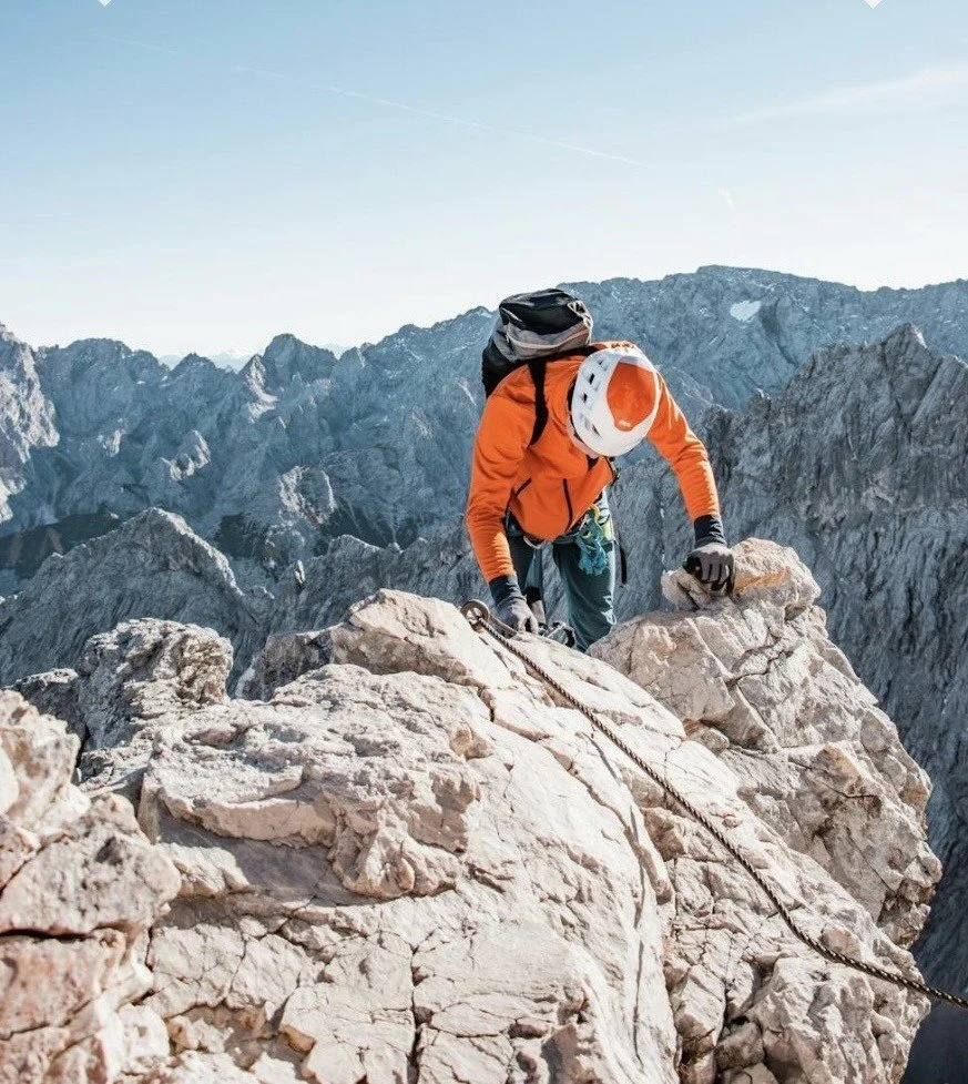 Alpiniste en escalade sur un rocher en montagne, portant un casque blanc, une veste orange, un sac à dos noir, et attaché à une corde d'escalade dans un paysage montagneux comme Chamonix