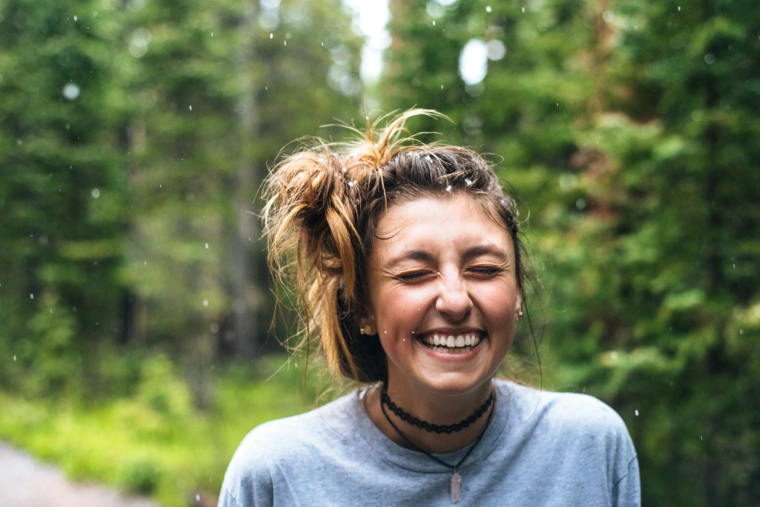 Une jeune femme souriante avec les yeux fermés, des cheveux attachés en un chignon désordonné, portant un t-shirt gris et un collier noir, dans un environnement naturel avec des arbres verts et de la pluie à Sallanches.