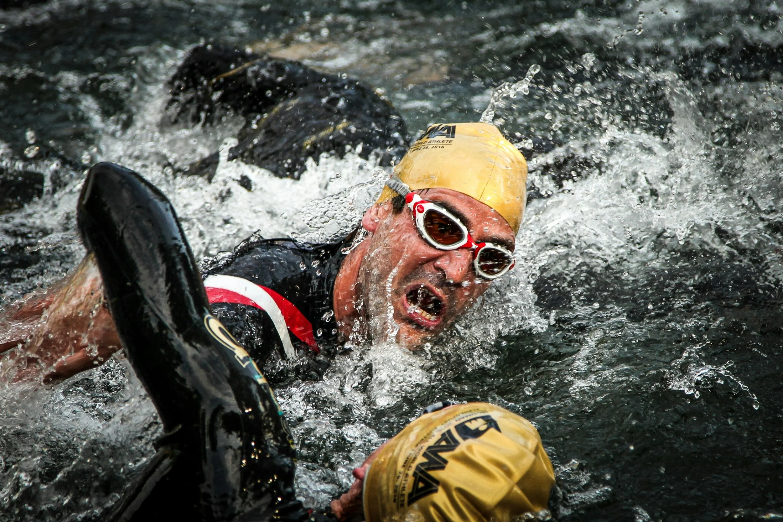 Un nageur triathlète portant une combinaison de natation, un bonnet jaune et des lunettes nage dans une rivière agitée, avec des éclaboussures d'eau tout autour dans le lac de Passy près de Sallanches