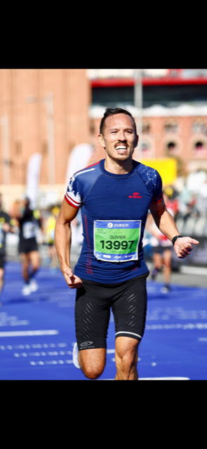 Un homme court un marathon, portant un t-shirt bleu avec un dossard tout en souriant. frère de Thierry Planté