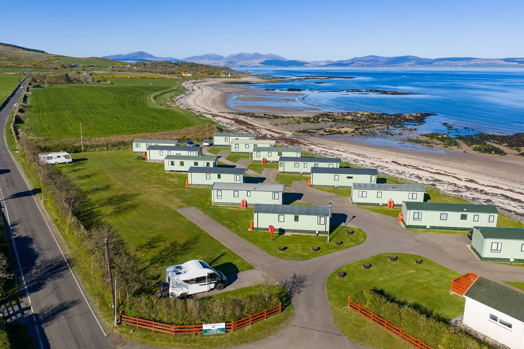 Aerial view of a coastal luxury holiday park with modern static caravans on a green lawn, overlooking a beach and calm ocean, with mountains in the distance.