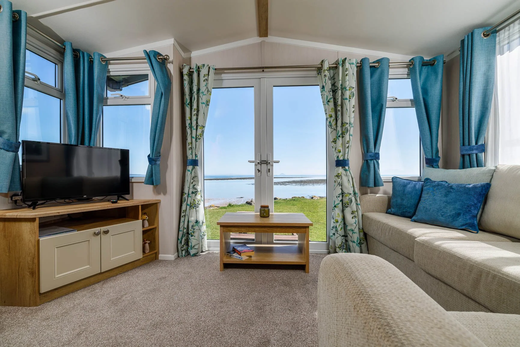 Living room with beige sofa and blue pillows facing large glass door and windows with blue curtains, ocean view with sky and grassy area outside, small coffee table with a jar in front of the door, TV on a wooden stand to the left, and beige carpeted floor.