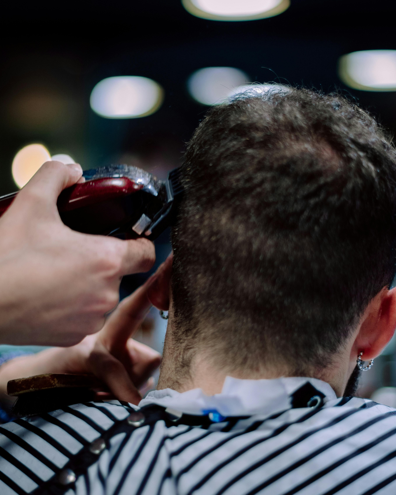 A person is getting a haircut using clippers, seen from behind, with short hair, in a barber shop with blurred lights in the background.