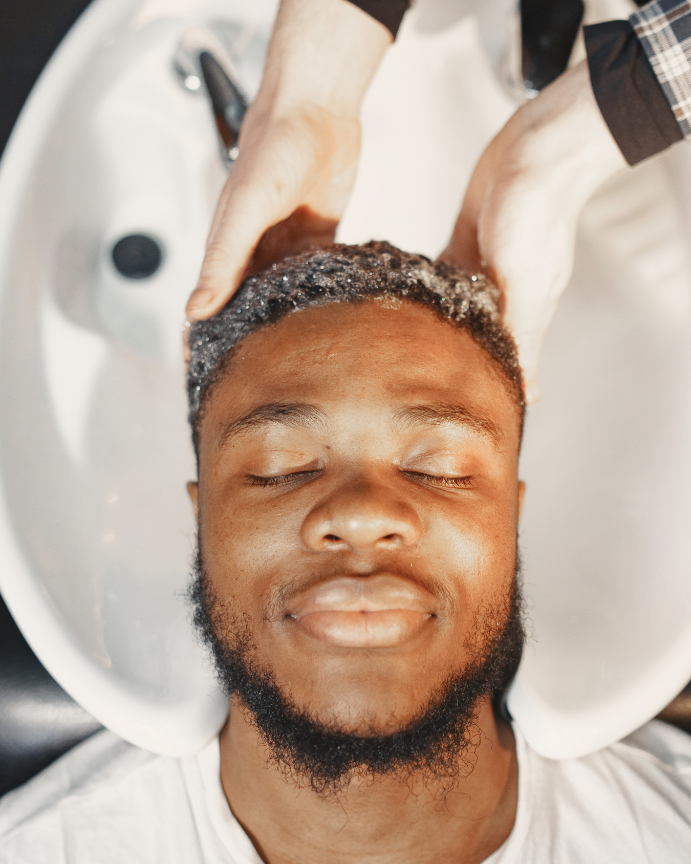 A man with closed eyes having his hair washed in a salon sink by a stylist wearing gloves.