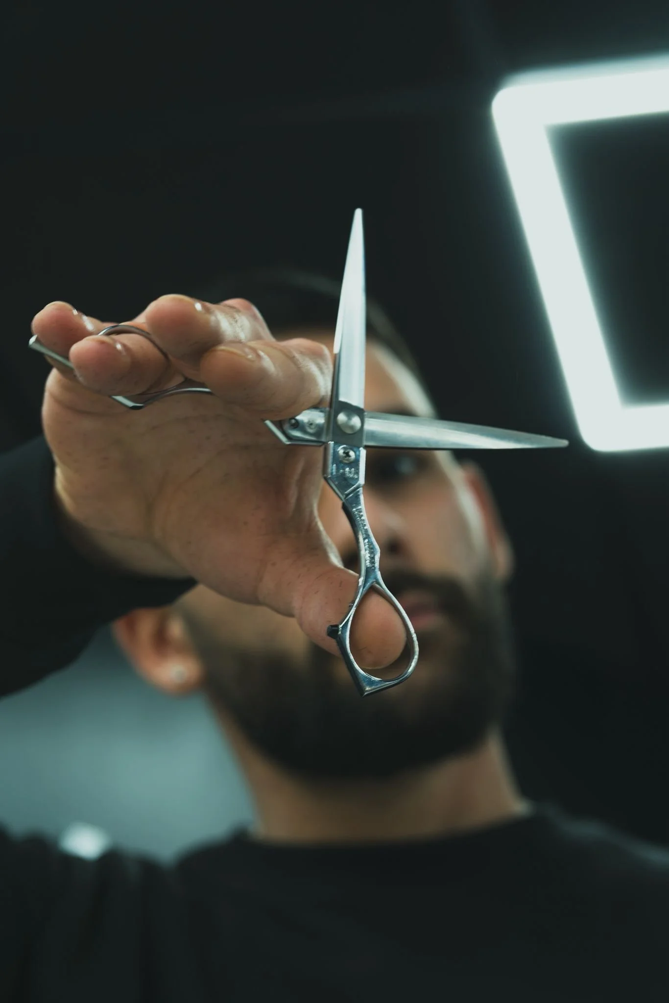 A person holding a pair of scissors near their ear, as part of a haircut or grooming process, with a bright light source and dark background.