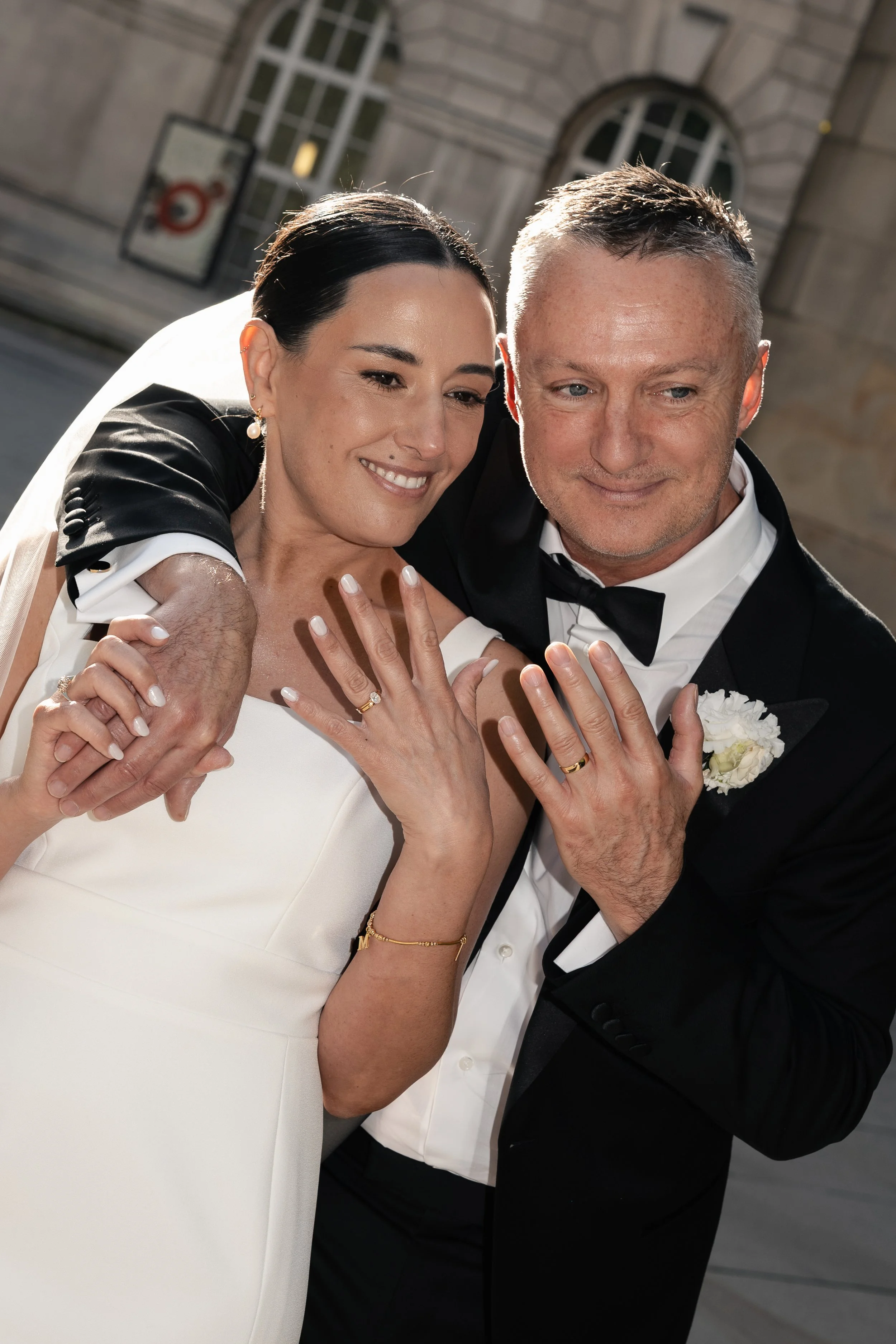 A newlywed couple dressed in formal wedding attire showing their rings while holding hands, standing outside a building with large windows.