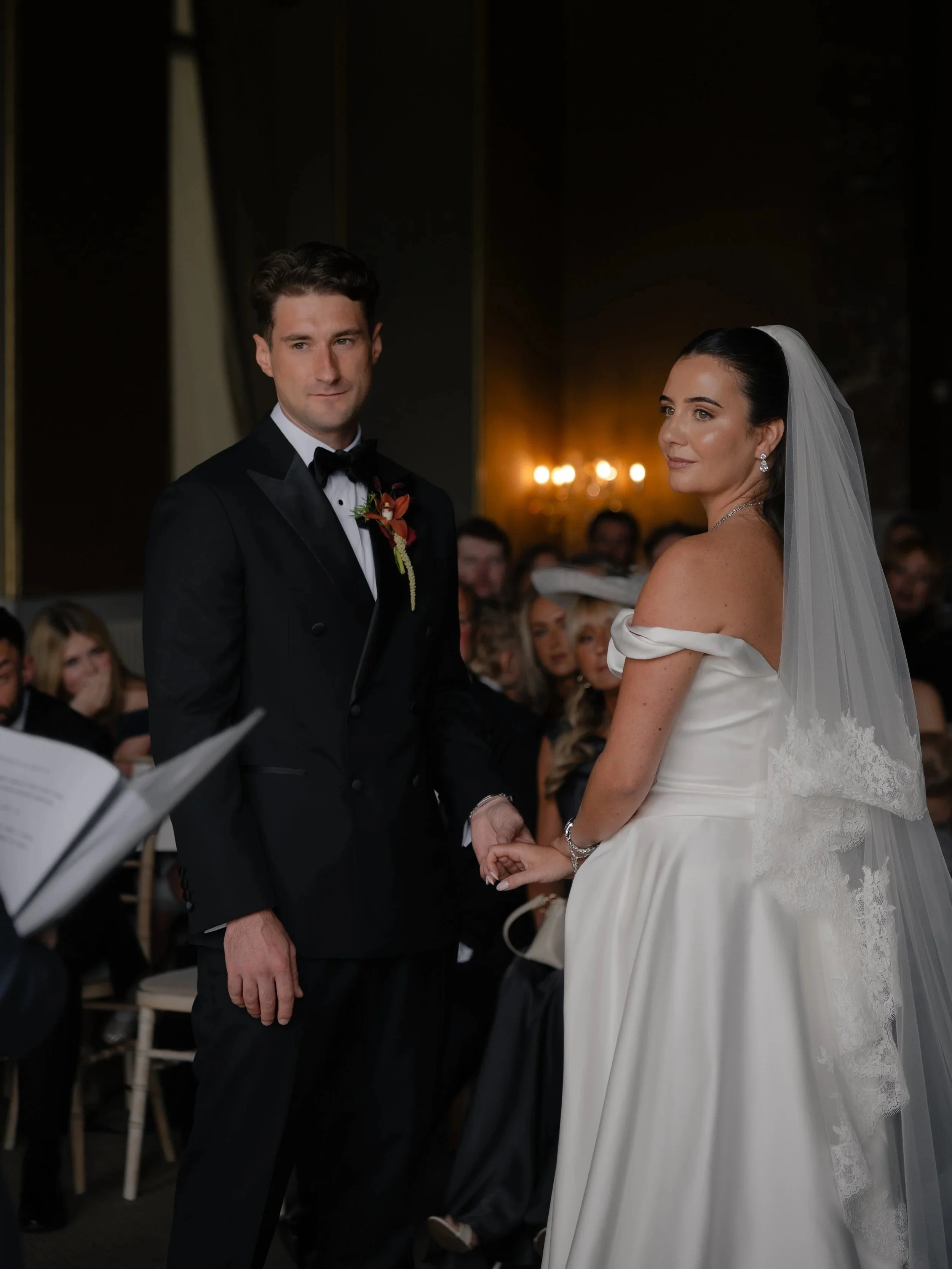 A bride and groom holding hands during their wedding ceremony indoors, with guests seated in the background.