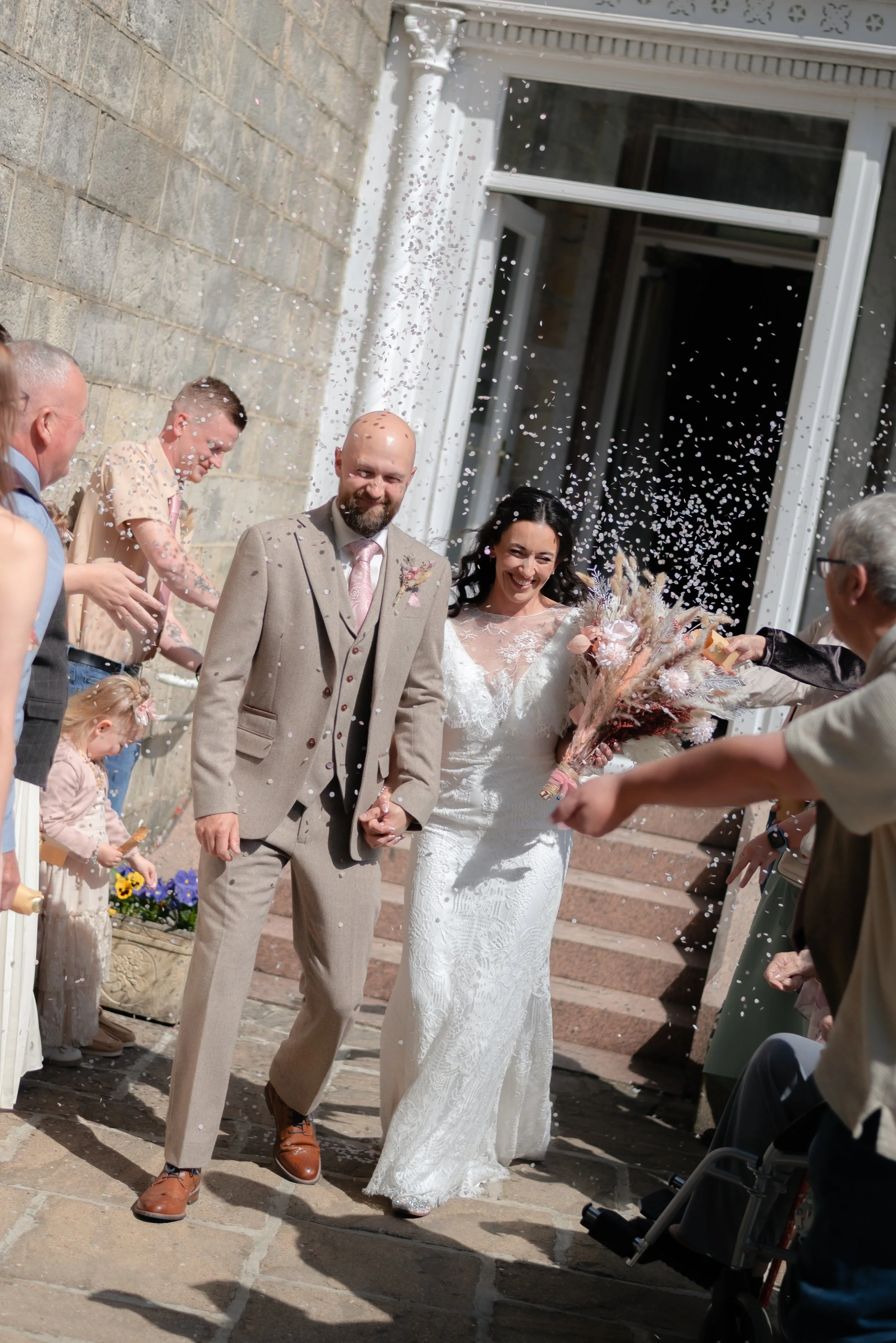 A newlywed couple walking outdoors, surrounded by guests throwing confetti, in front of a building with large windows. The groom wears a beige suit and pink tie, while the bride wears a white lace wedding dress and holds a bouquet of flowers.
