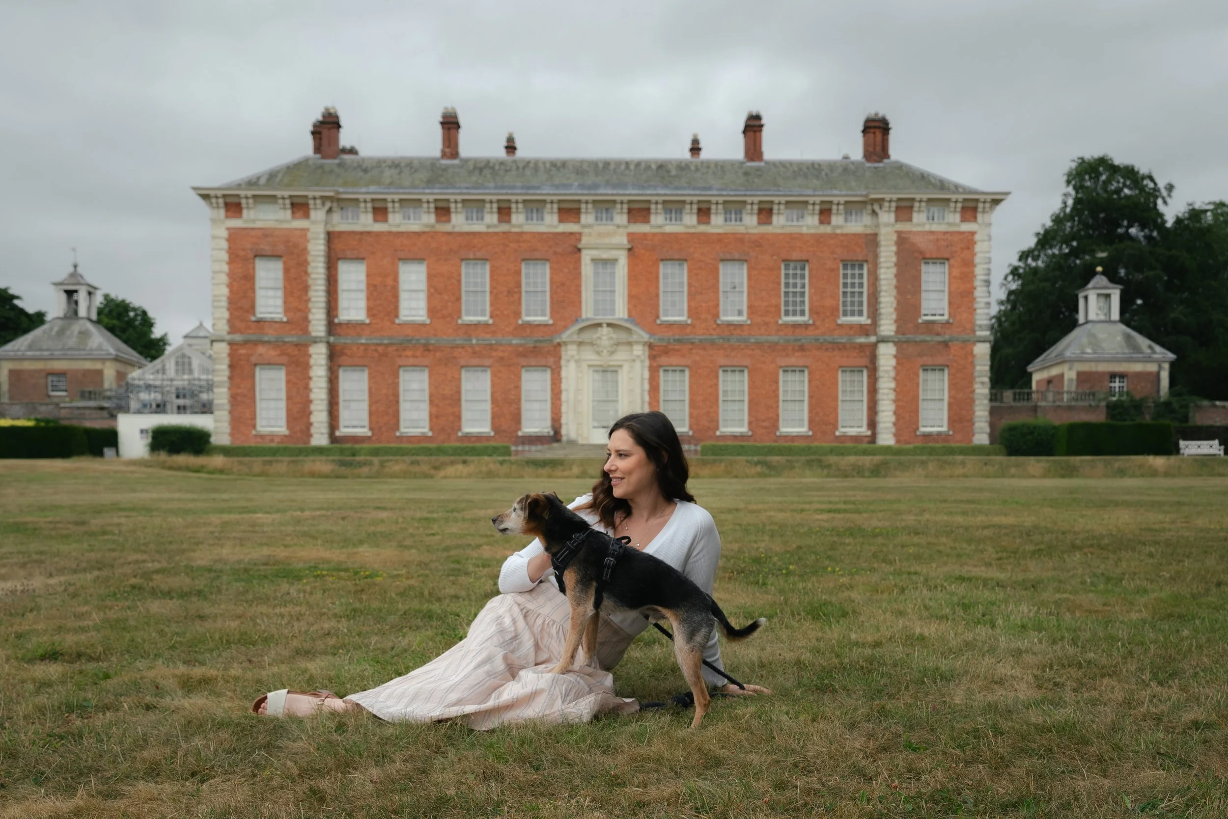 A woman sitting on the grass holding a dog in front of a large historic brick building with multiple chimneys and smaller buildings on either side, on a cloudy day.