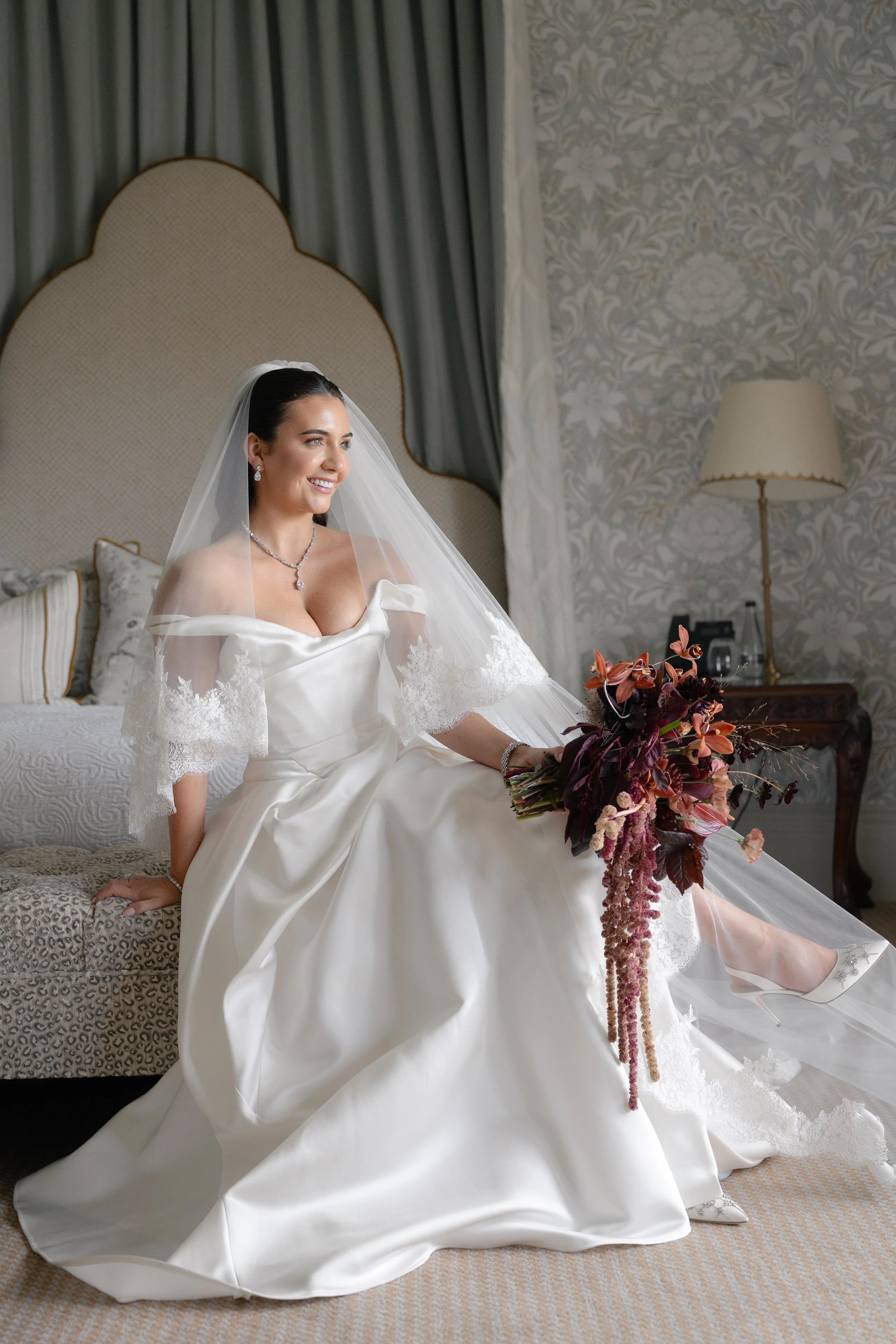 A smiling bride wearing a white wedding gown with lace embroidery on the sleeves and a veil, sitting on a patterned bench in a hotel room, holding a dark red and pink flower bouquet.