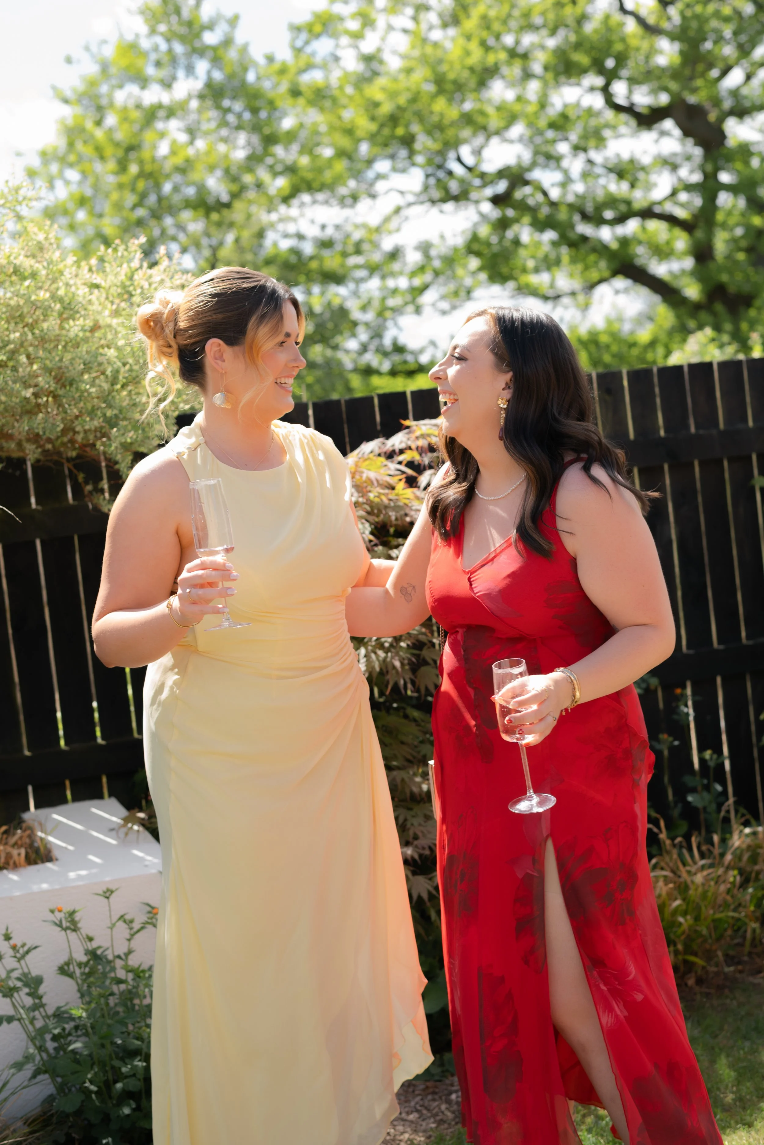 Two women in elegant dresses smiling and talking outdoors on a sunny day, holding champagne glasses, with green trees and a black wooden fence in the background.
