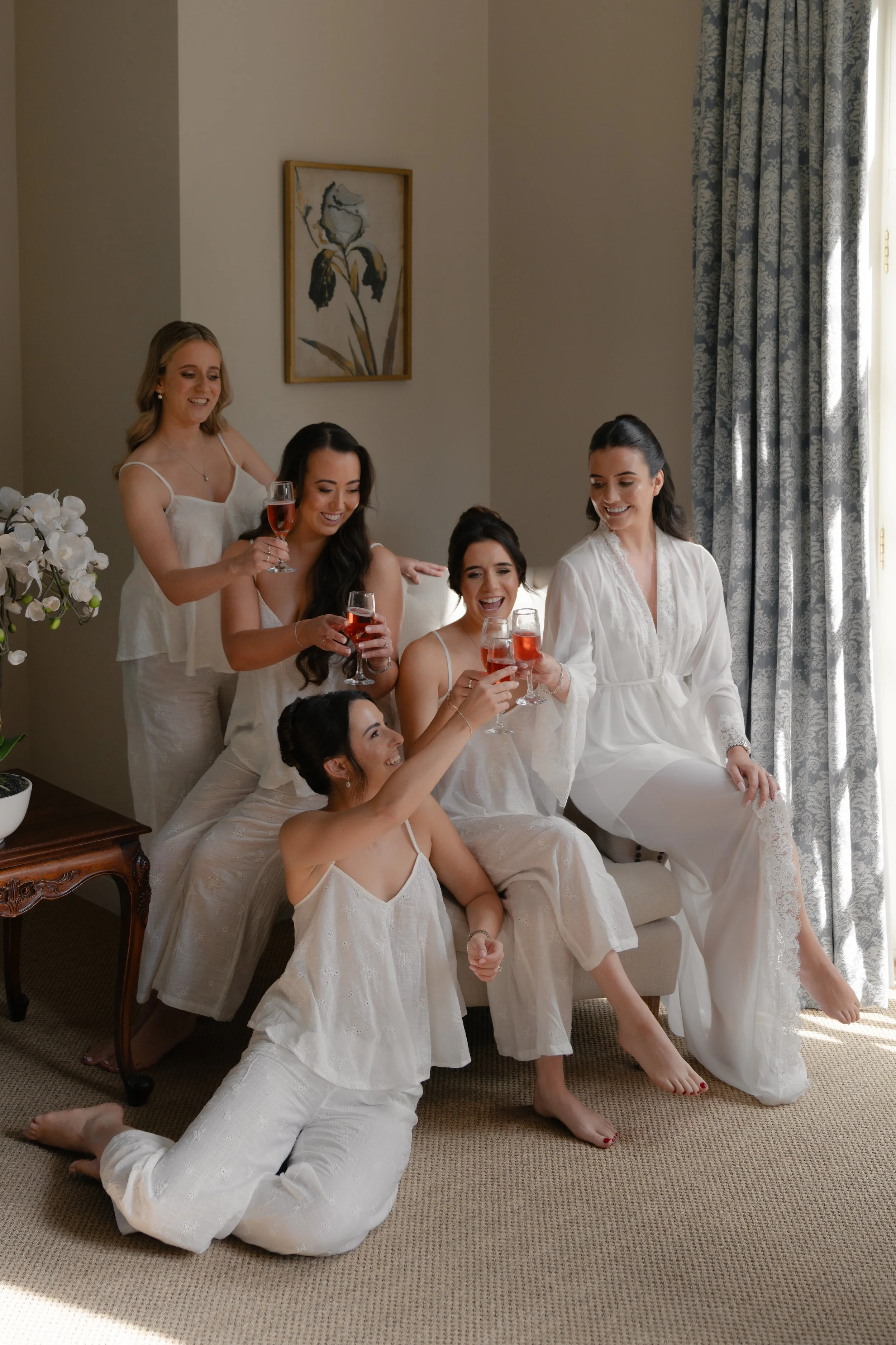 Group of six women in white pajamas celebrating with drinks in a living room with beige walls, a window with blue patterned curtains, and a framed floral artwork.