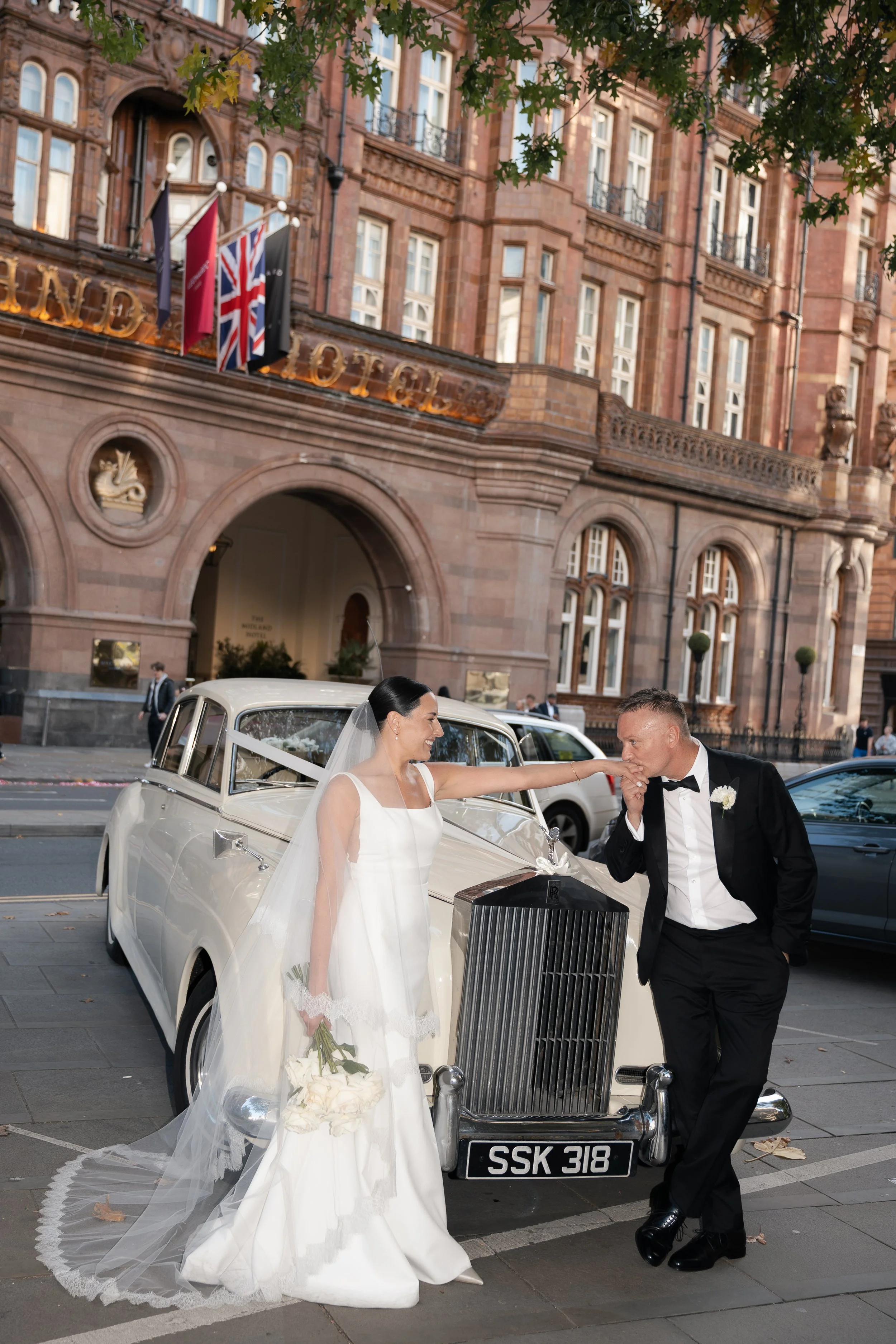 A bride in a white wedding dress with a veil holding a bouquet, and a groom in a black tuxedo holding a hand to his mouth, standing next to a vintage white car with a black grille outside a red brick building. The bride is smiling and the groom is lo