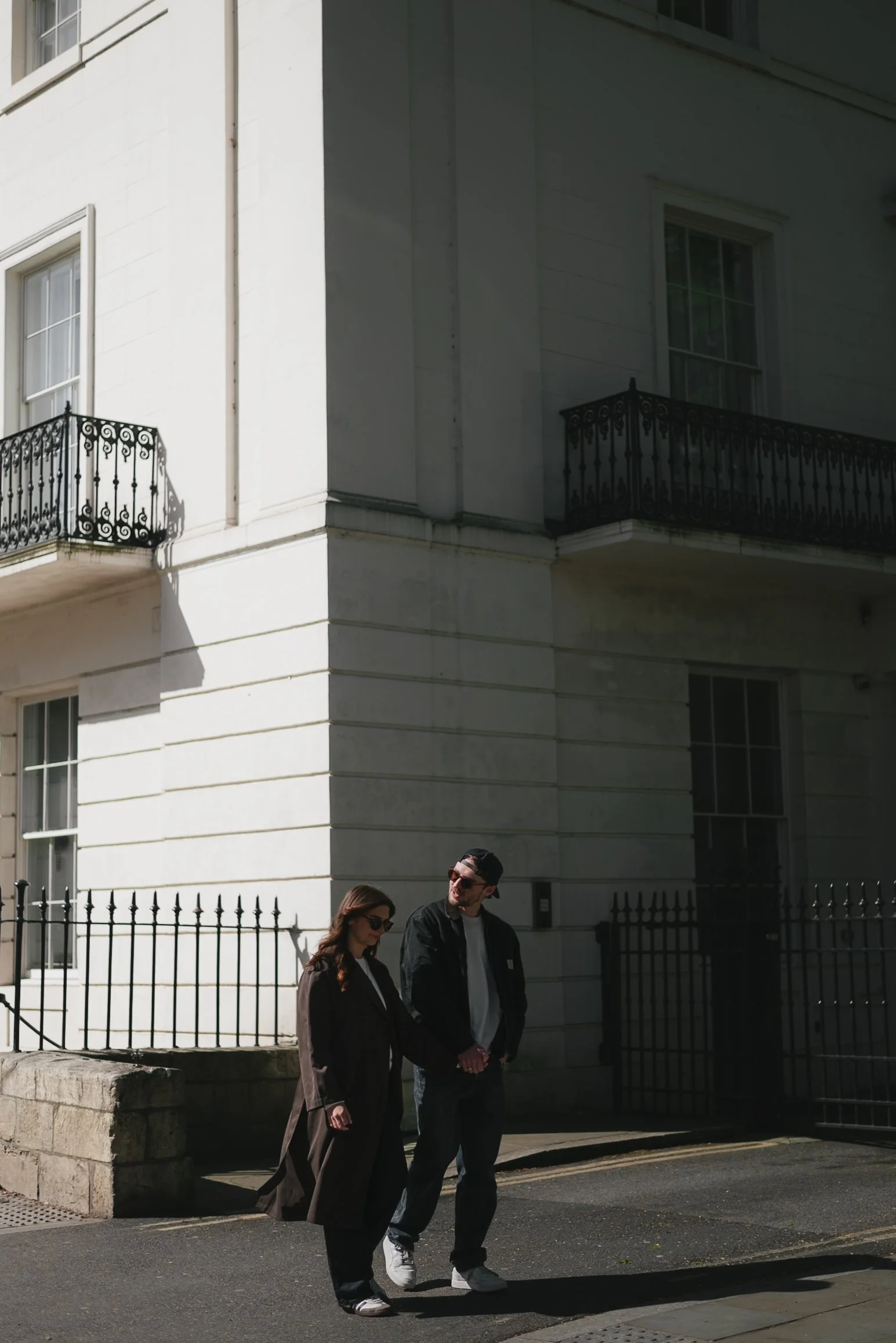 A man and woman walking on a city sidewalk in front of a large white building with black wrought-iron balconies and a decorative fence. The woman is wearing sunglasses, a long coat, and dark pants, while the man is wearing sunglasses, a dark jacket, and white sneakers.