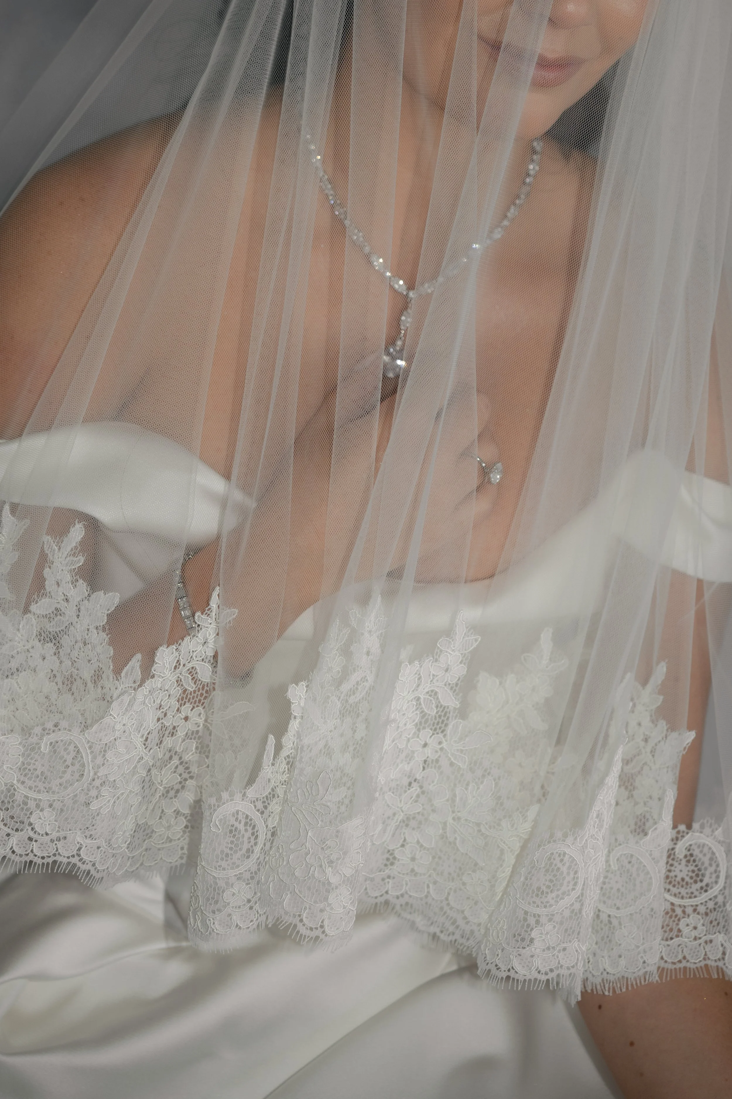 A bride wearing a white wedding dress with lace detailing, champagne-colored lipstick, and a pearl necklace, partially obscured by a sheer white veil.