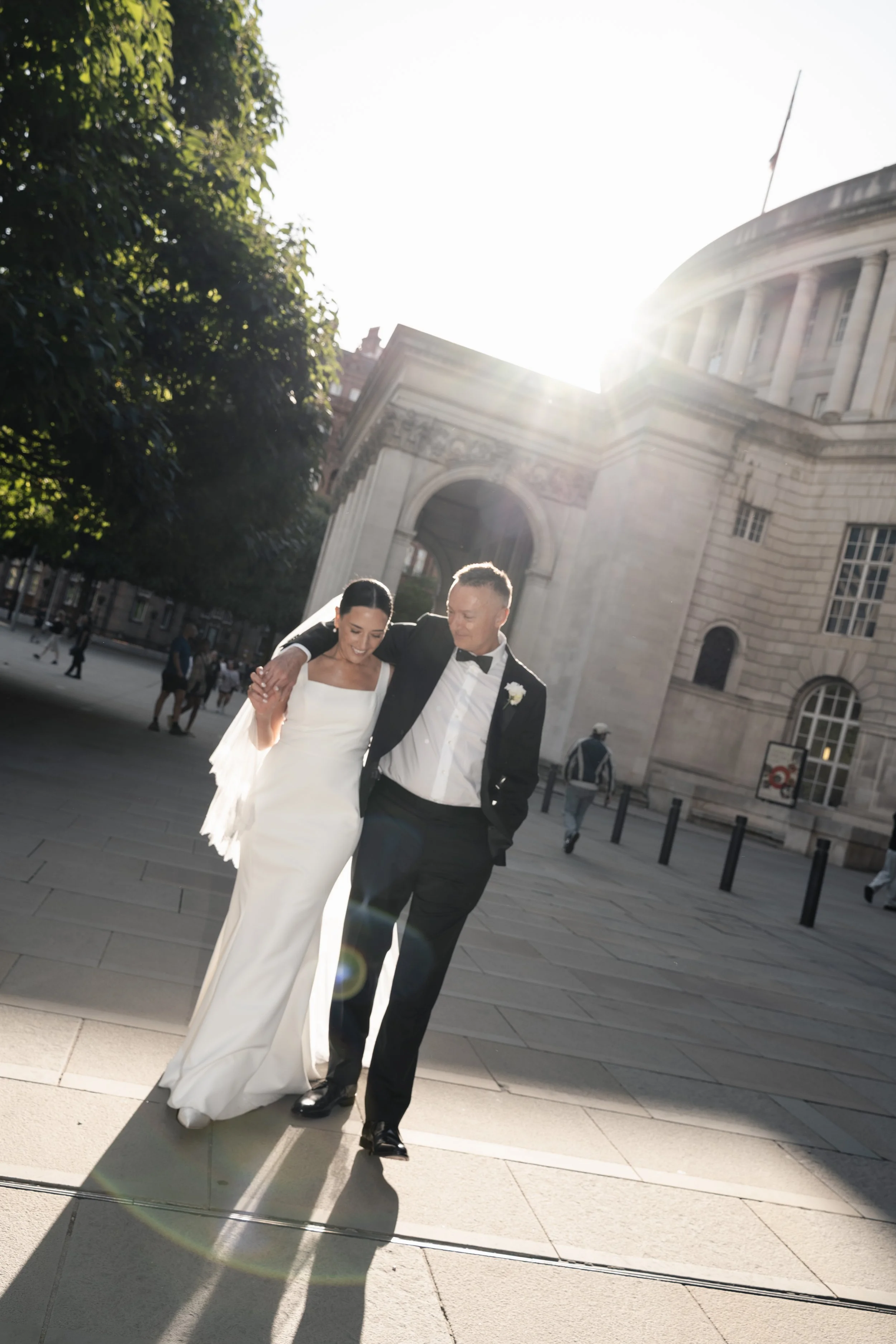 A bride and groom walking arm in arm outside in front of a large stone building with columns and an archway, illuminated by bright sunlight.