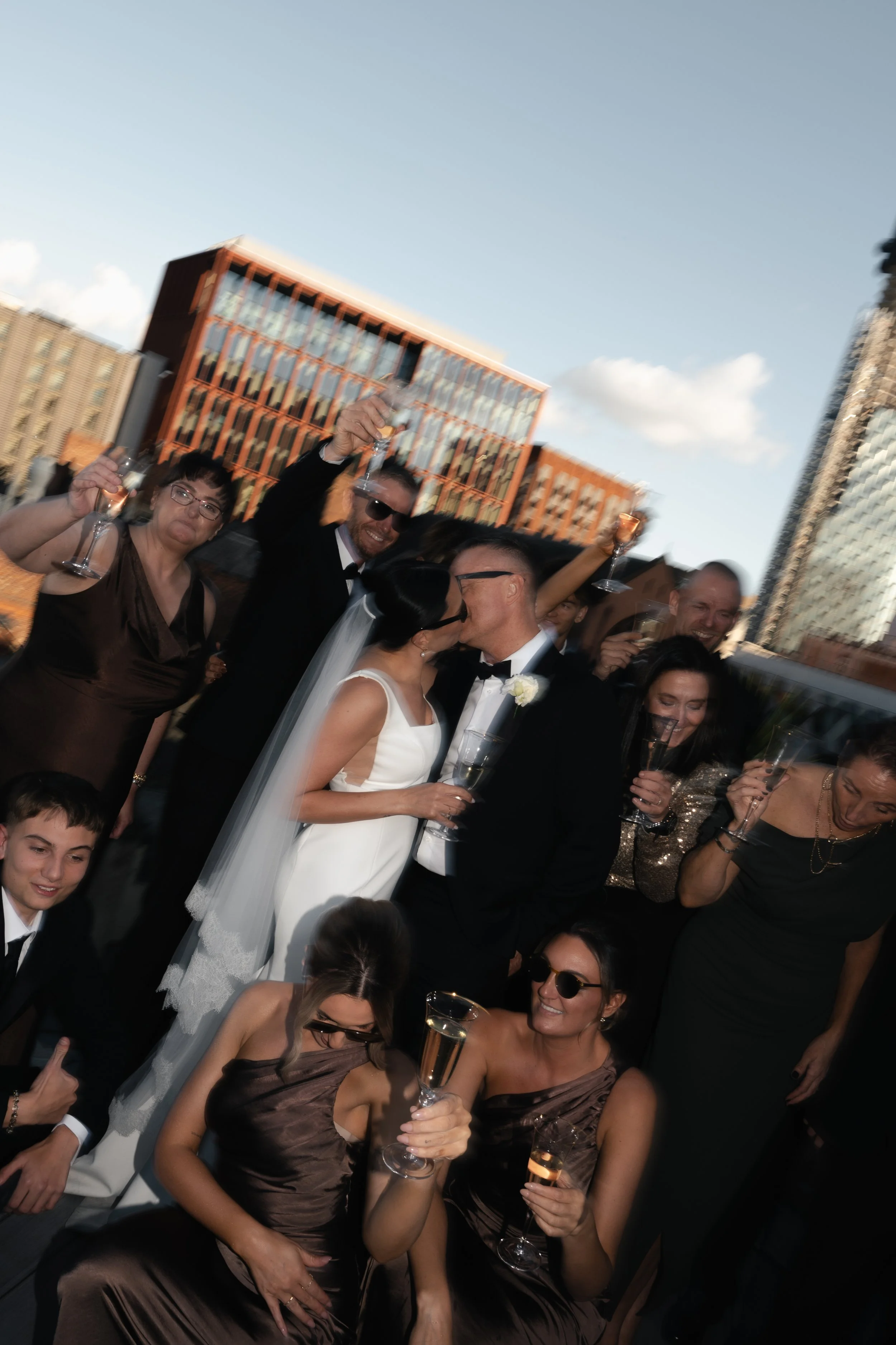 Group of people celebrating at an outdoor party, wearing formal attire, holding glasses of champagne, with city buildings in the background.