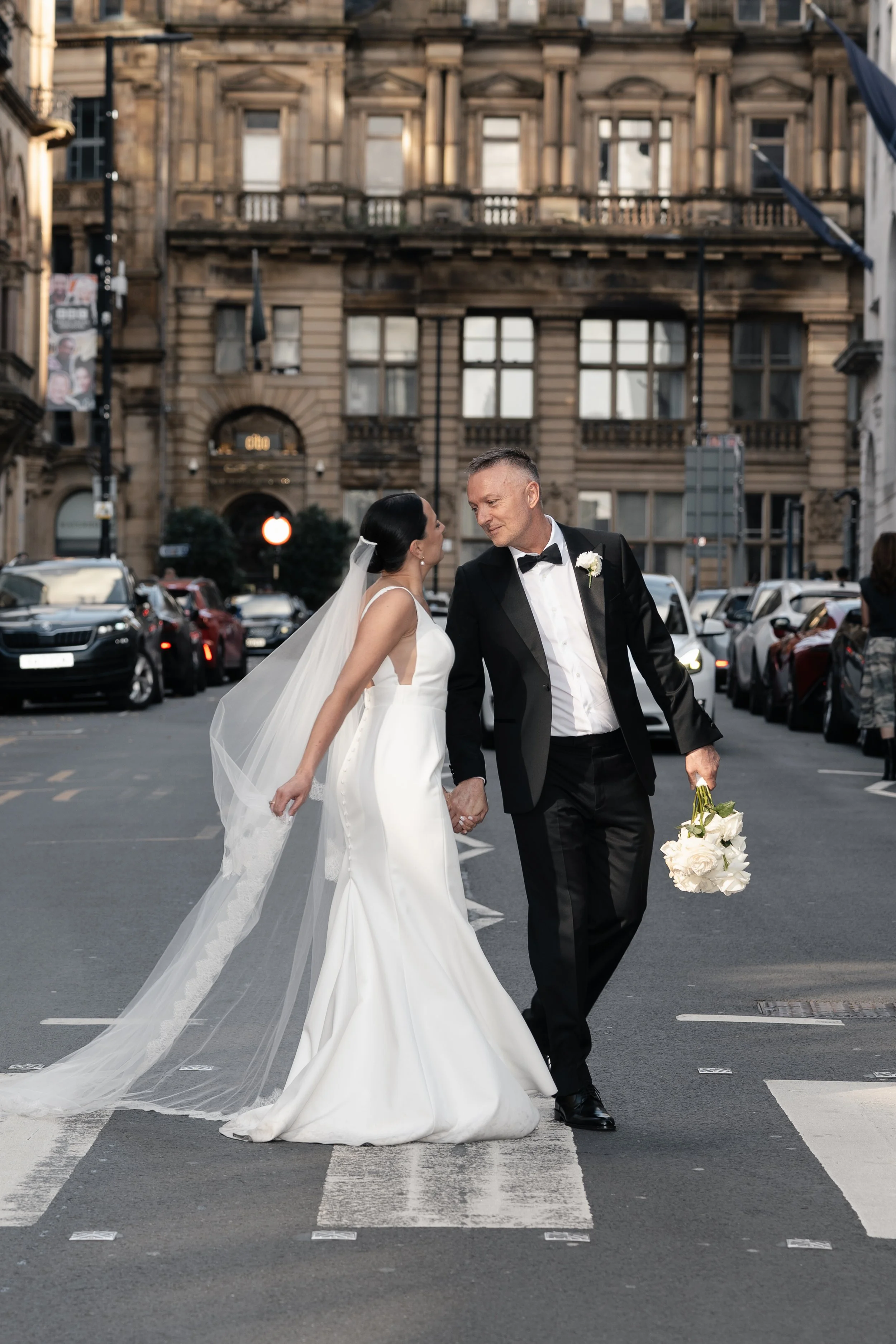 A bride and groom dancing on a city street at sunset. The bride is wearing a white wedding gown and veil, and the groom is in a black tuxedo with a bow tie, holding a bouquet of white roses.