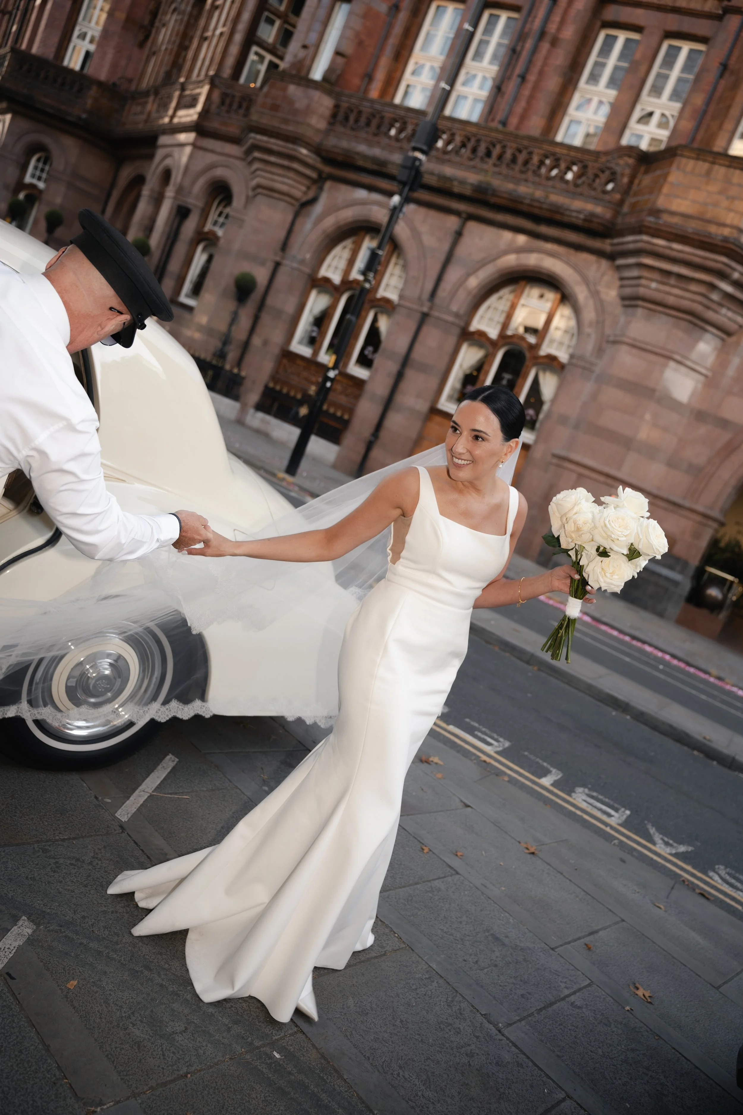 A bride in a white wedding gown holding a bouquet of white roses shaking hands with a man in a white shirt and black hat in front of a historic brick building.