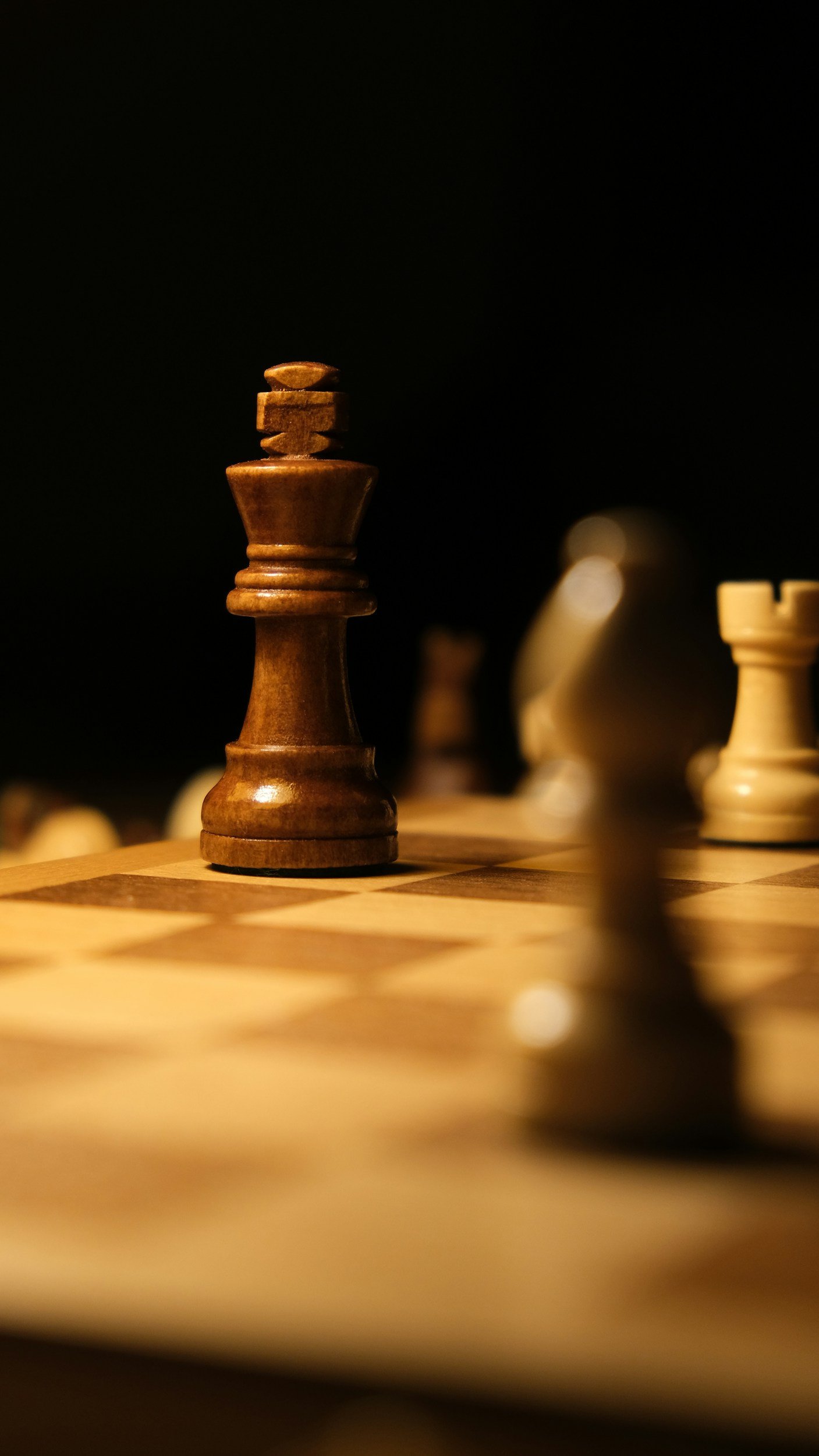 A chess king piece in focus on a chessboard, with other pieces blurred in the background, against a dark background.