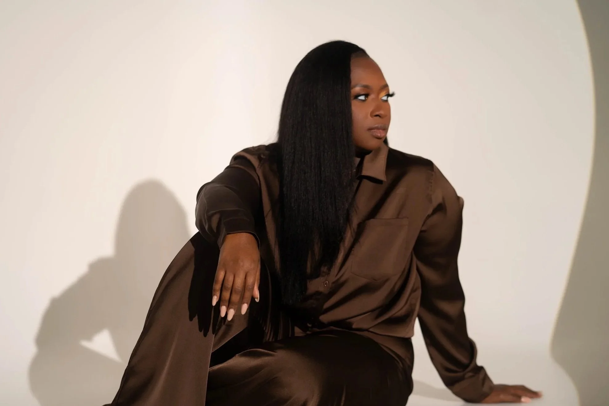 A woman with long black hair, sitting on the floor with her back against a wall. She is wearing a brown outfit, and her left hand rests on her knee. The background is plain white.