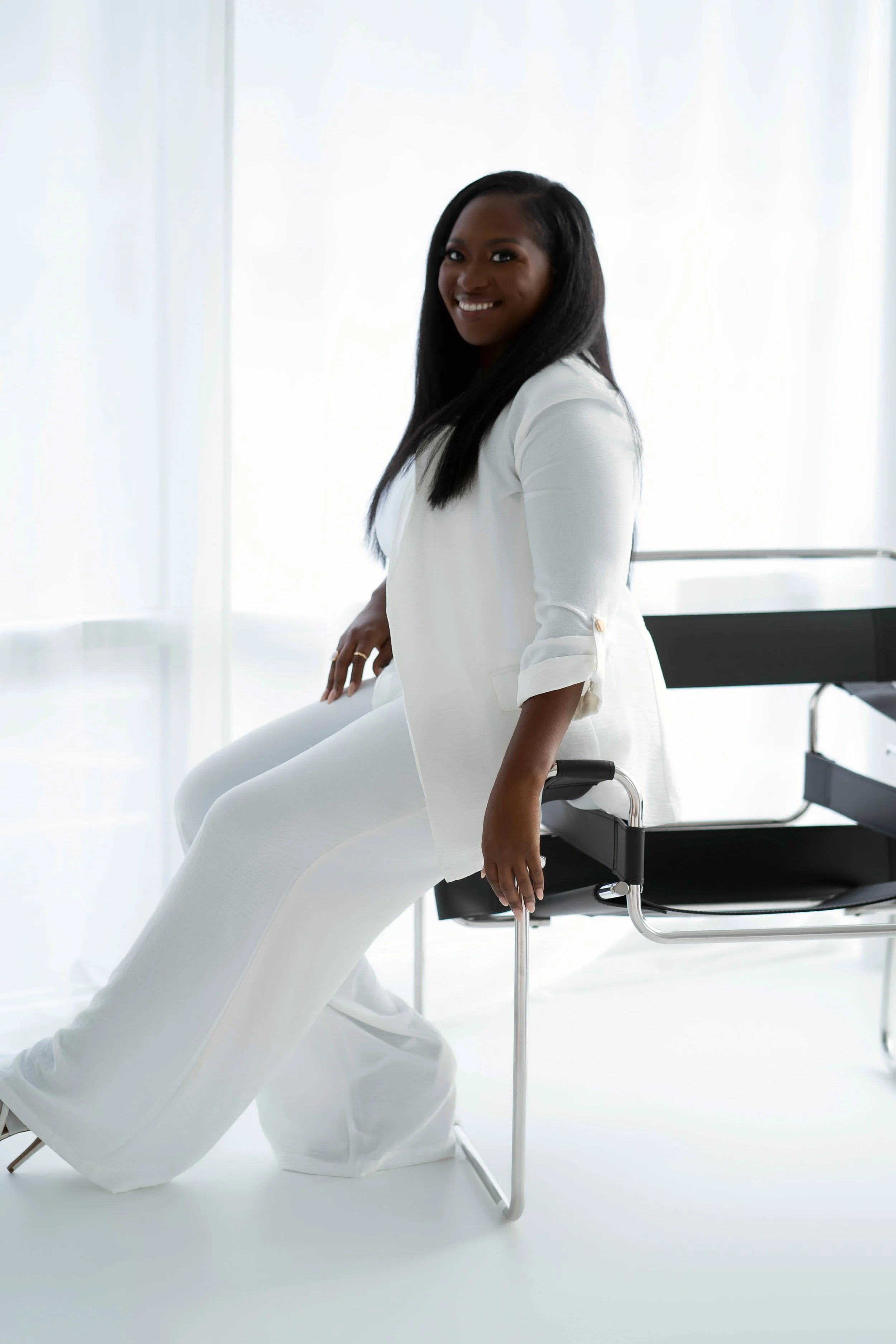 A woman sitting on a modern white medical chair in a bright room, smiling at the camera, dressed in white clothing.