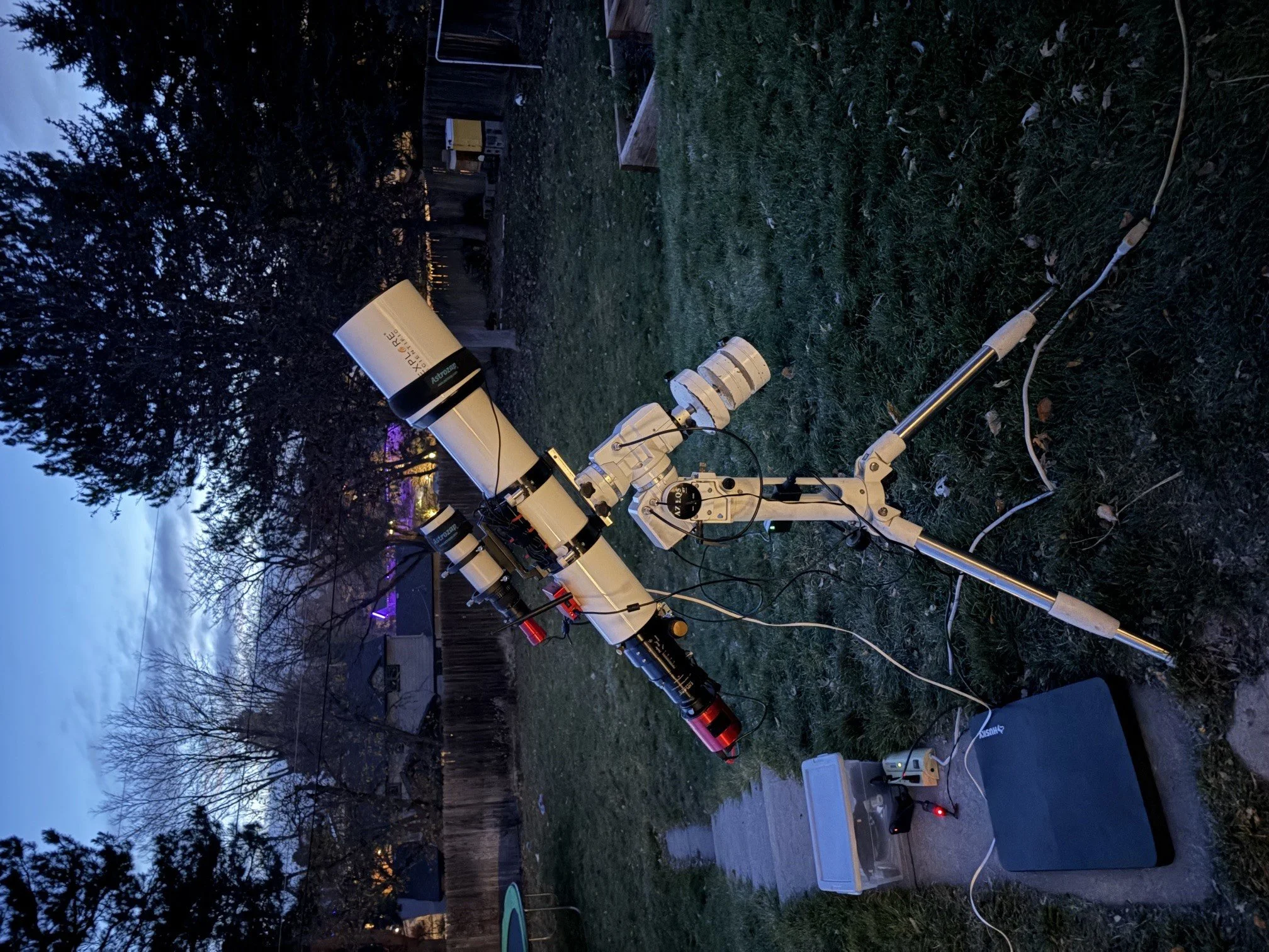 A telescope setup outdoors in a backyard at dusk, with equipment arranged on the grass and various electronic devices and boxes nearby.
