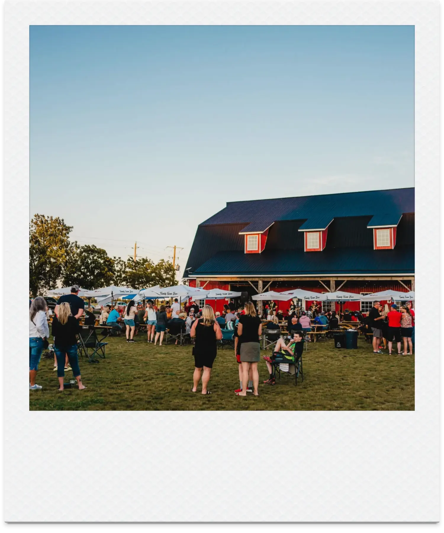 A large outdoor gathering with people sitting and standing on grass, under umbrellas, near a red barn-like building with a black roof and small dormer windows, during what appears to be late afternoon or evening at a Chatham, Ontario event