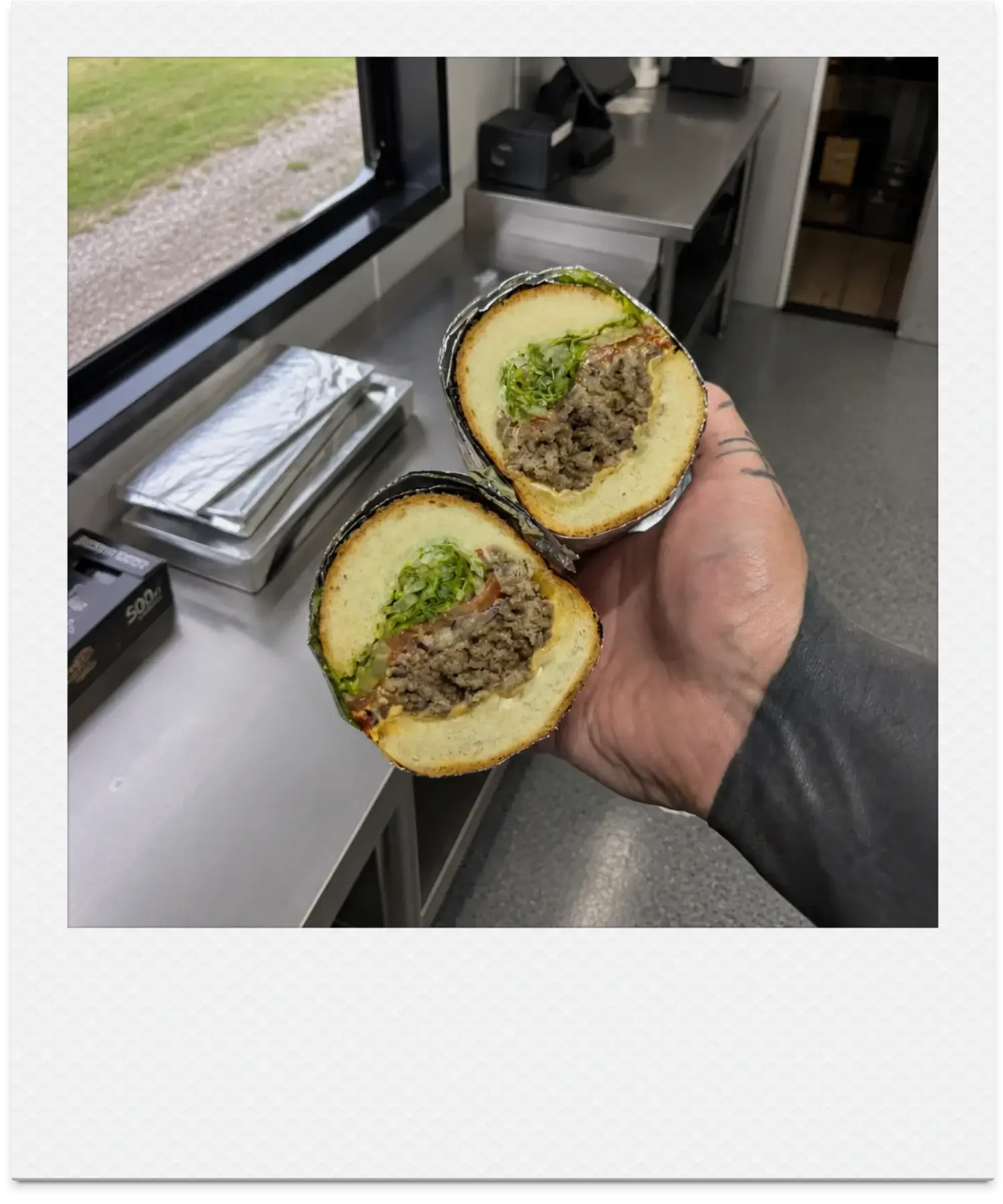Person holding a sandwich containing shredded beef, lettuce, and green onions inside a bun, in a kitchen setting.