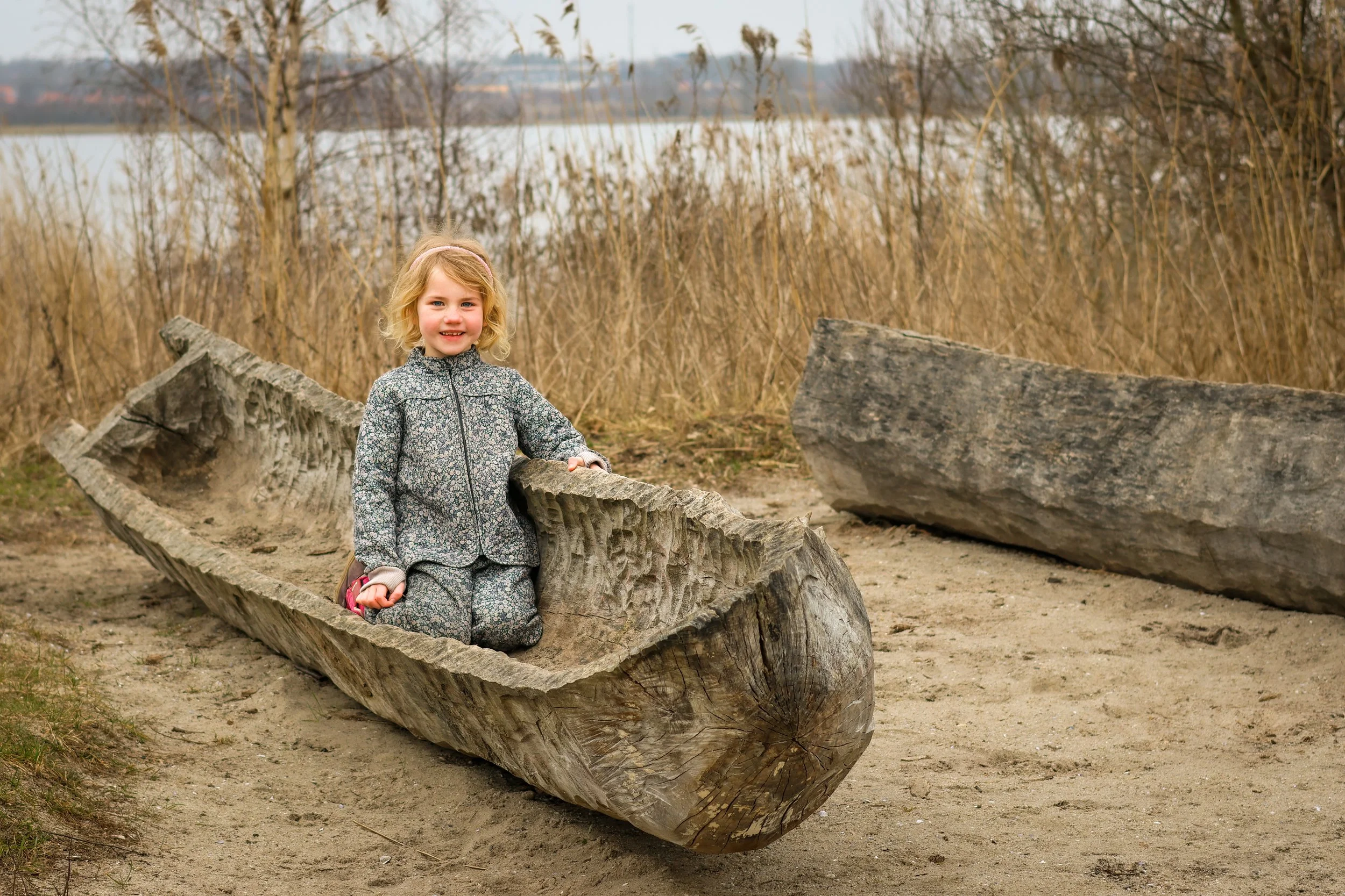 En ung pige med blond hår, grå tøj, sidder i en gammel træbåd, der ligger på en sandbund ved en vandkant, omgivet af tørre græsser og buske, med en sø eller flod i baggrunden.