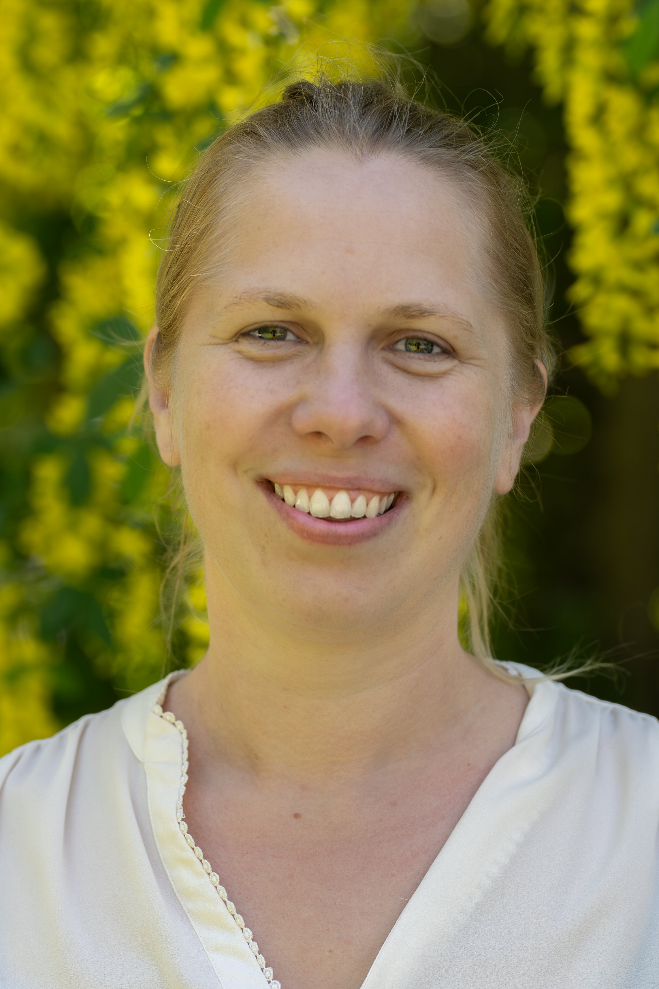 En smilende kvinde med blond hår, iført en hvid top, står foran en grøn og gul baggrund af blomster