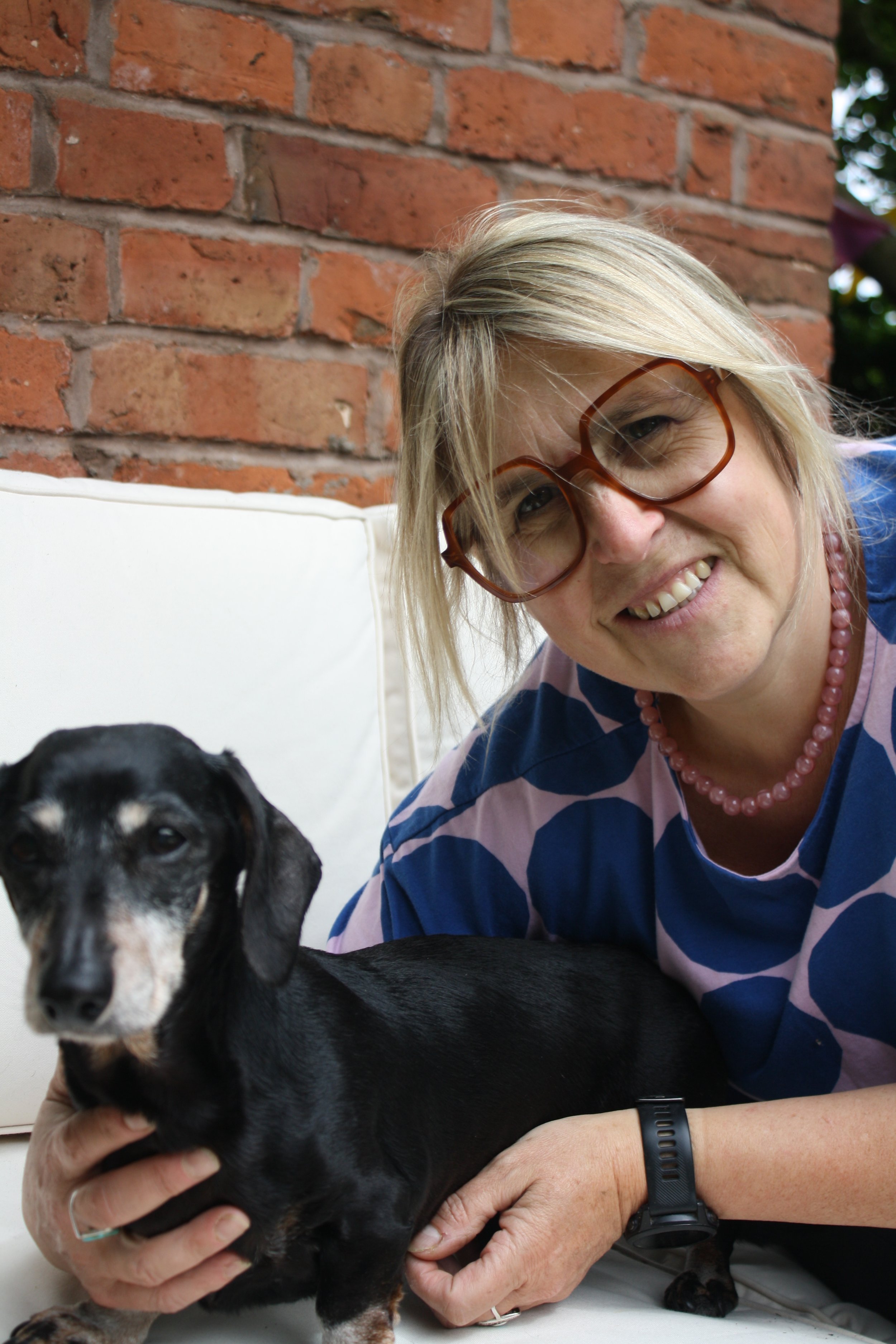 A smiling woman with blonde hair, wearing glasses, a pink pearl necklace, and a striped shirt, is holding a black and tan puppy in her hands. They are sitting outdoors against a brick wall background.