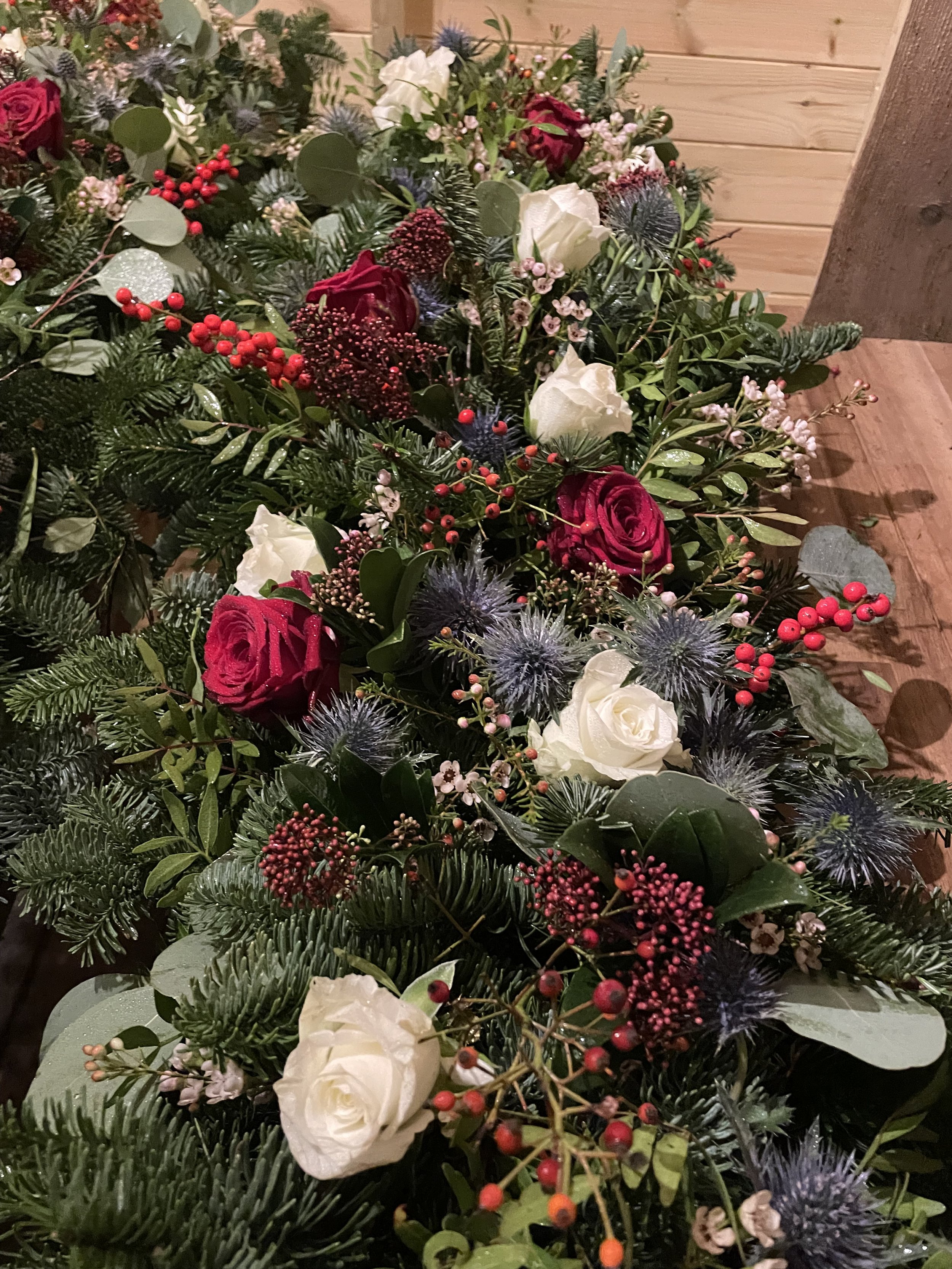 Close-up view of a Christmas wreath with red roses, white roses, berries, eucalyptus, pine branches, and blue thistle-like flowers, placed on a wooden floor.