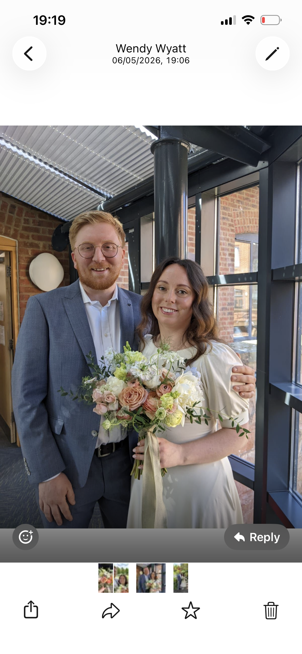A man and woman standing together indoors, with the man in a grey checkered blazer and glasses, the woman holding a bouquet of flowers, both smiling at the camera, with a brick wall and large windows in the background.