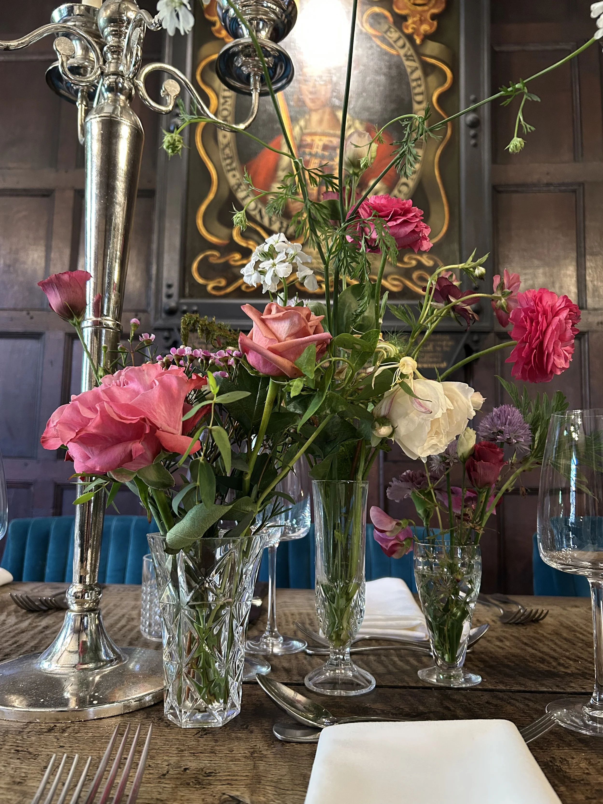 A dining table with several glass vases holding pink, white, and purple flowers. A silver candelabra with black candlesticks is on the table. In the background, there is a large portrait of a man in historical clothing with a dark wood-paneled wall.