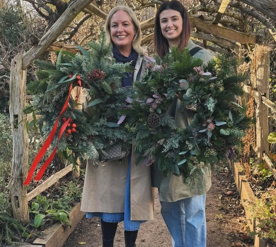 Two women standing outdoors in a garden holding large holiday wreaths decorated with evergreen branches and red berries.