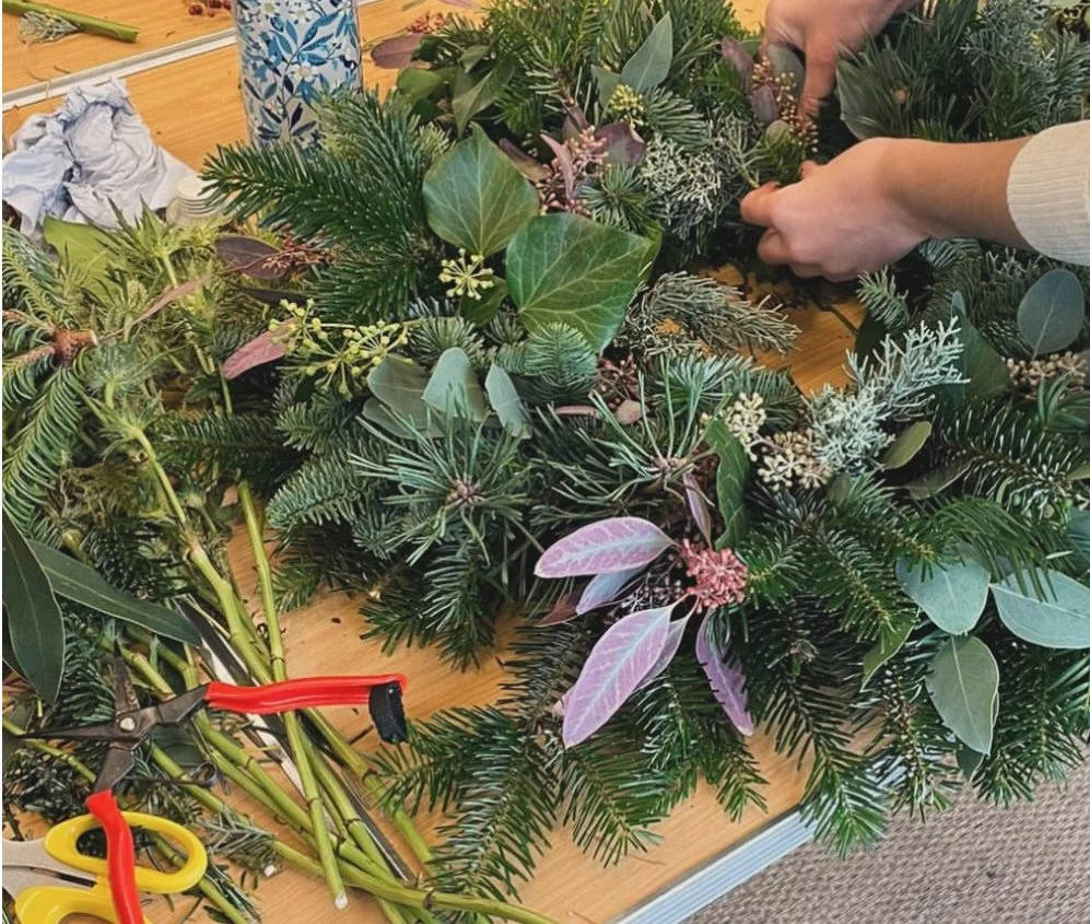 Hands arranging a Christmas wreath made with various types of evergreen and purple-tinted leaves on a wooden table.