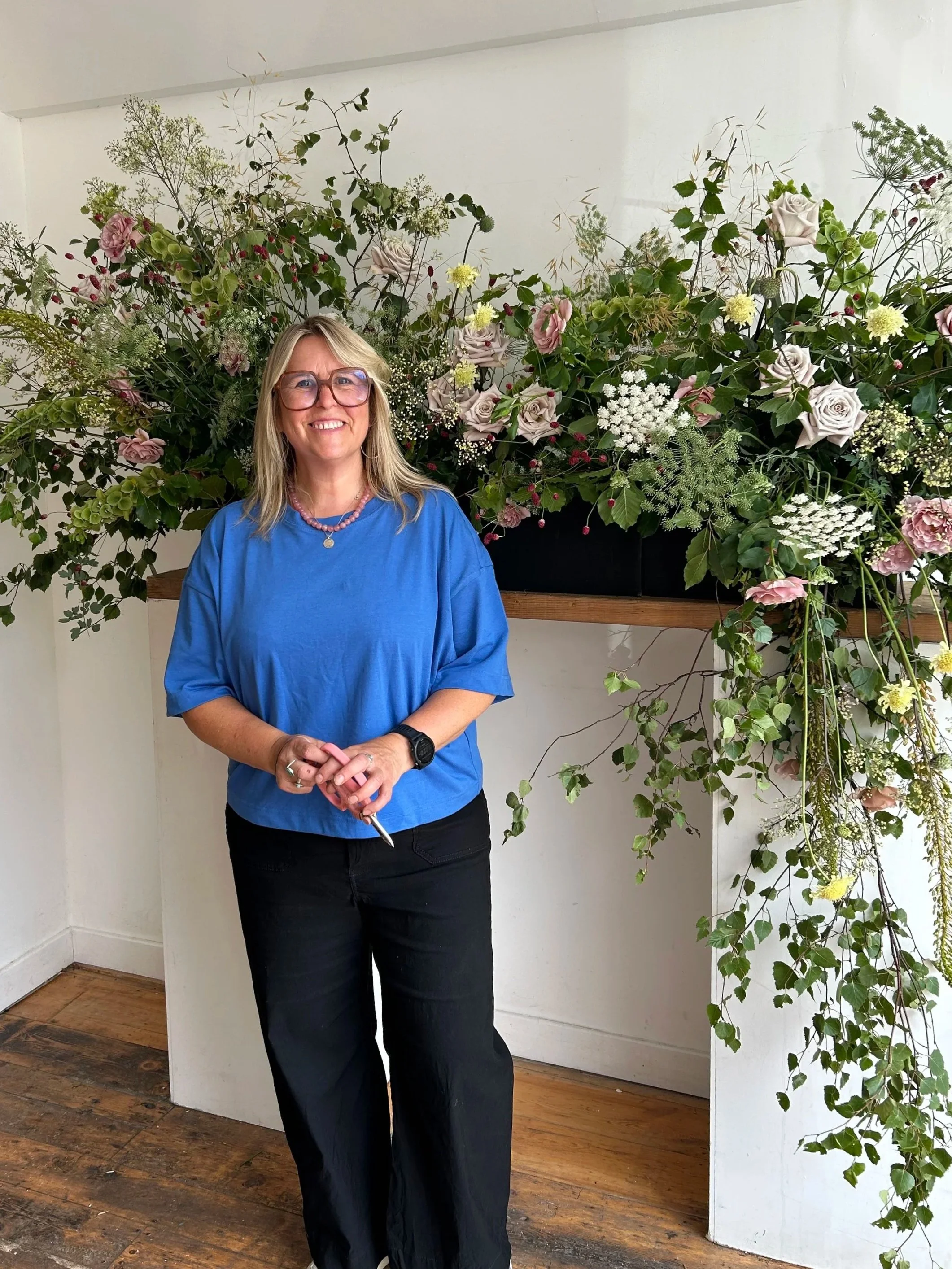 A woman with blonde hair and glasses smiling, wearing a blue top and black pants, standing in front of a floral arrangement with green leaves and various flowers, including roses, in an indoor setting with wooden flooring.